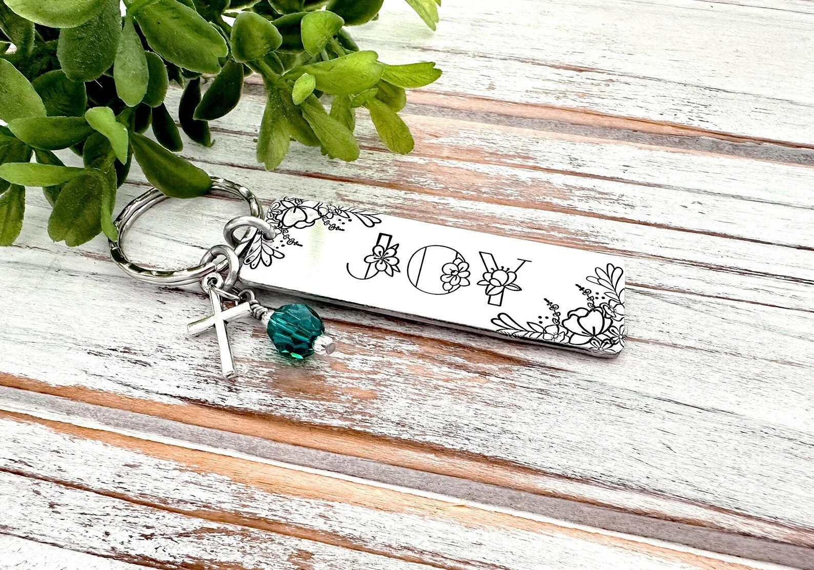 A decorative keychain with a silver cross charm, a teal bead, and a rectangular metal tag with floral patterns and the word 'LOVE' engraved, next to a leafy green plant on a white wooden surface.