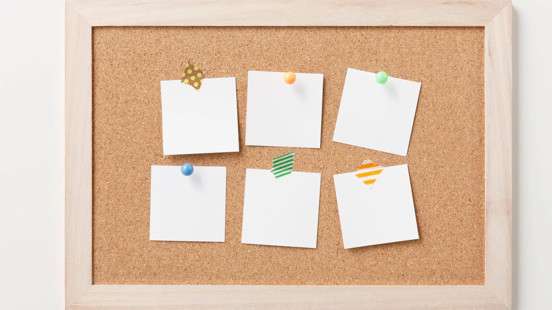 Cork bulletin board with six blank white papers pinned with colorful push pins and decorative tape.