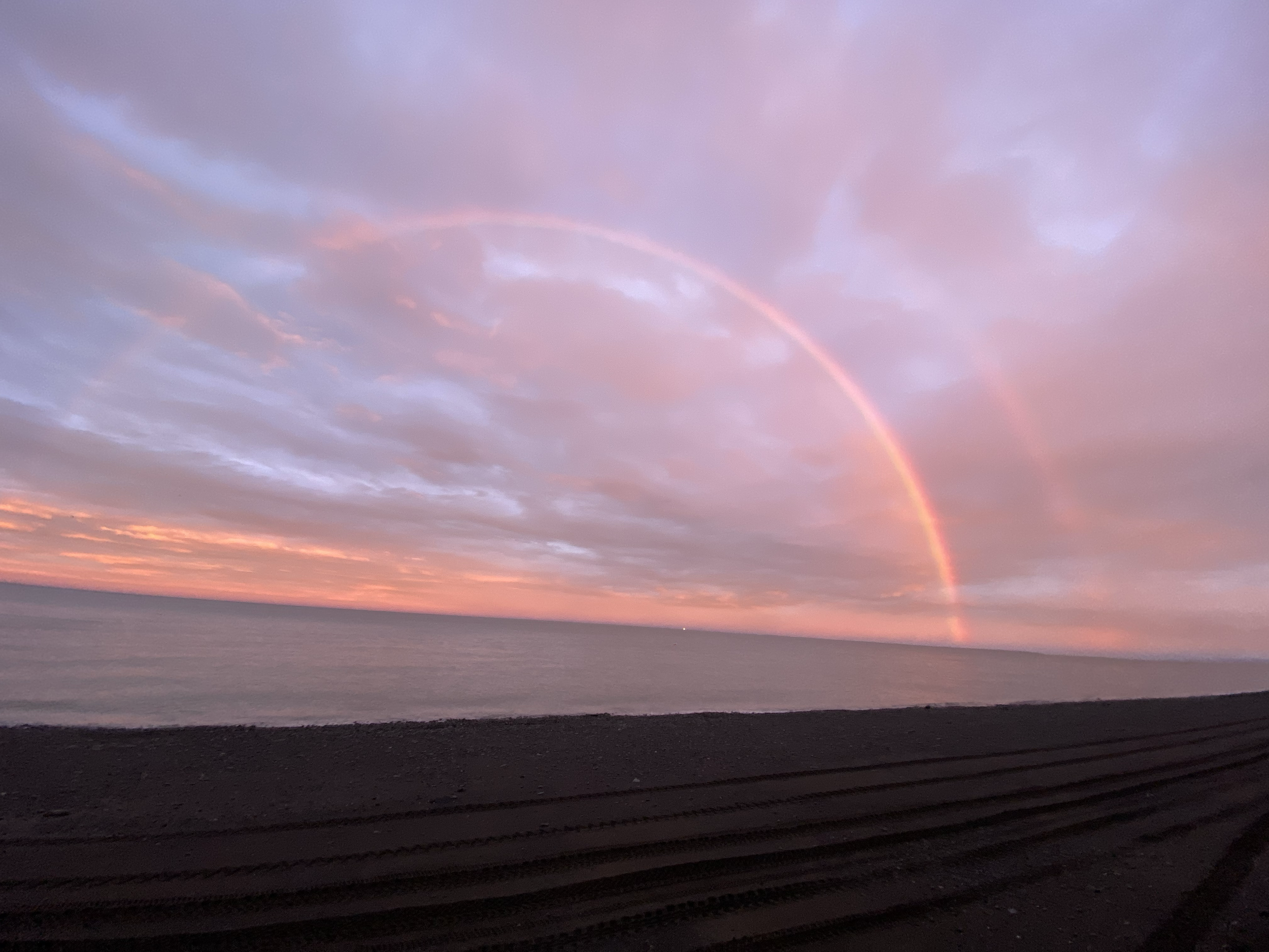 Pink and purple sunset over the ocean with a double rainbow in the cloudy sky and dark sandy beach in the foreground.