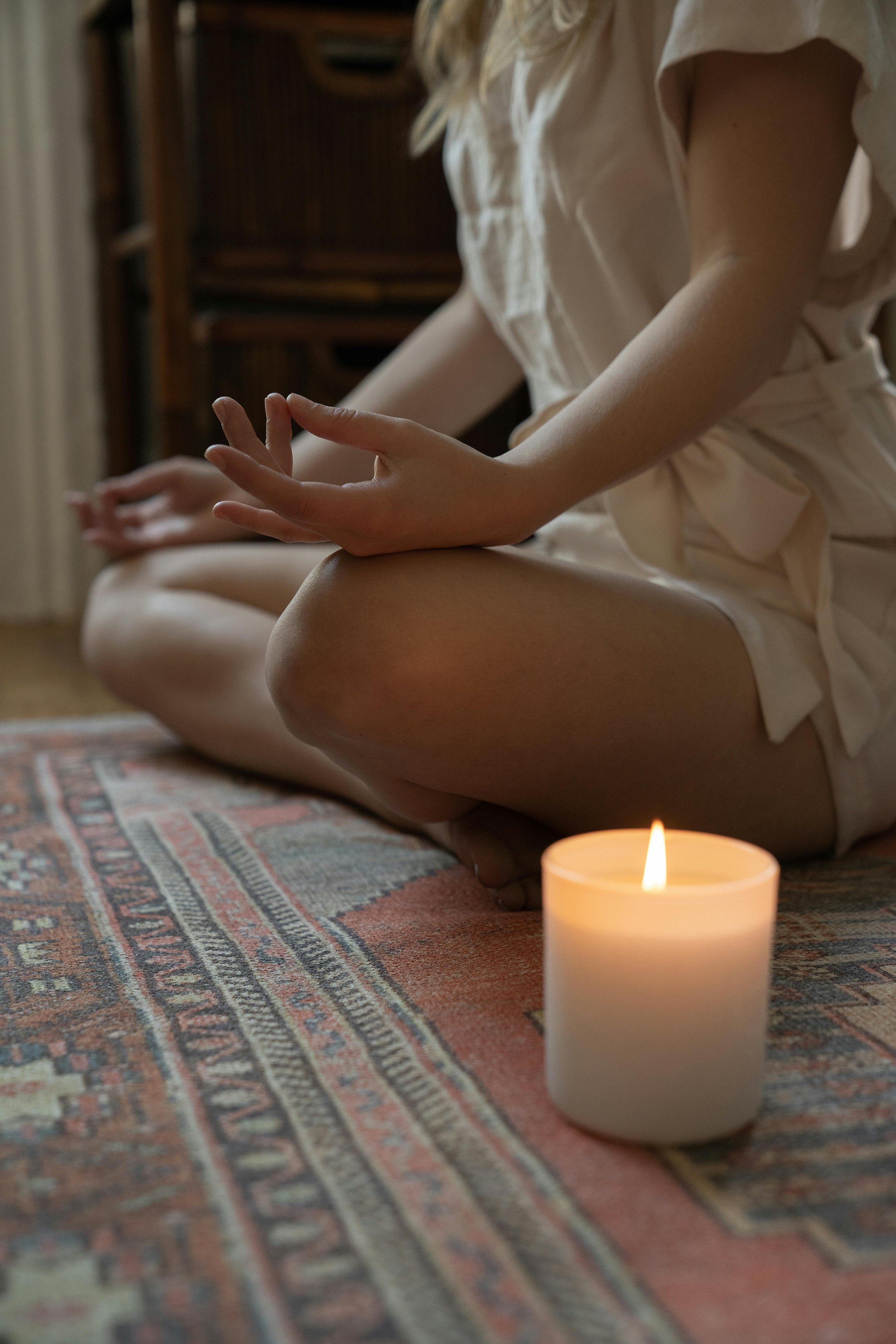 Person in light-colored clothing sitting on a patterned rug with a lit candle nearby, practicing meditation or mindfulness.