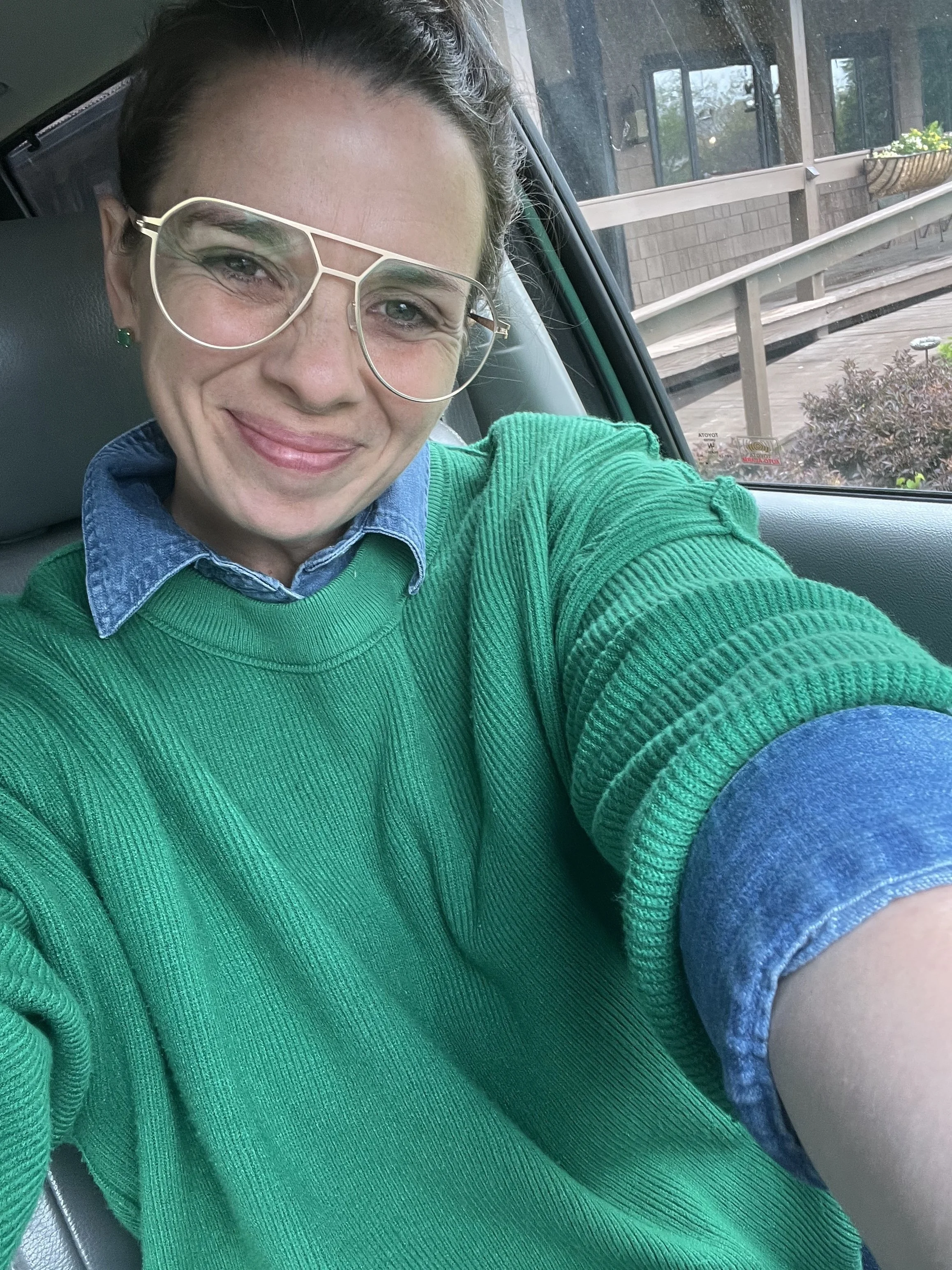 a selfie of Jenna wearing wire-rimmed glasses and a green sweater over a denim collared shirt