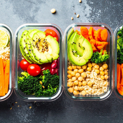 Rectangular glass containers full of healthy foods including avocado, tomatoes, broccoli, chickpeas, and carrots on a dark grey countertop