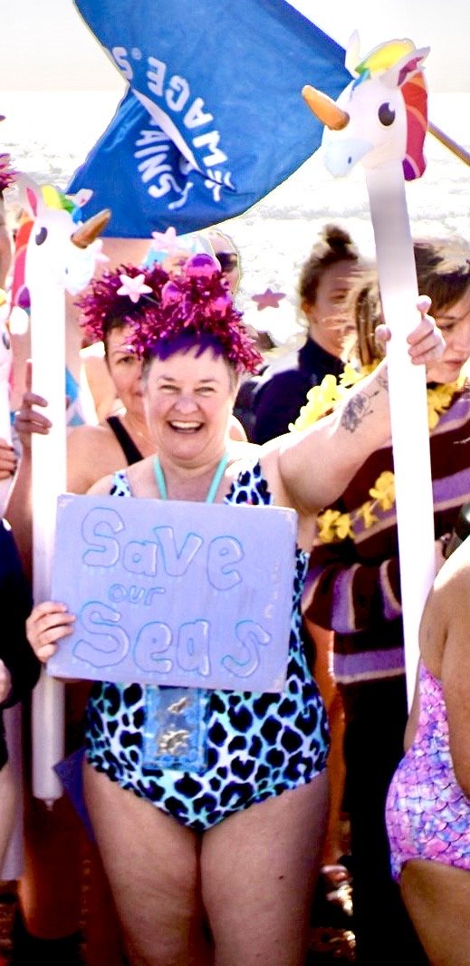 Zoe Carroll is a woman with short brown hair and a purple fringe. She wears a blue leopard print swimming costume, and is holding a banner that says Save Our Seas in blue paint, and is holding an inflatable unicorn.