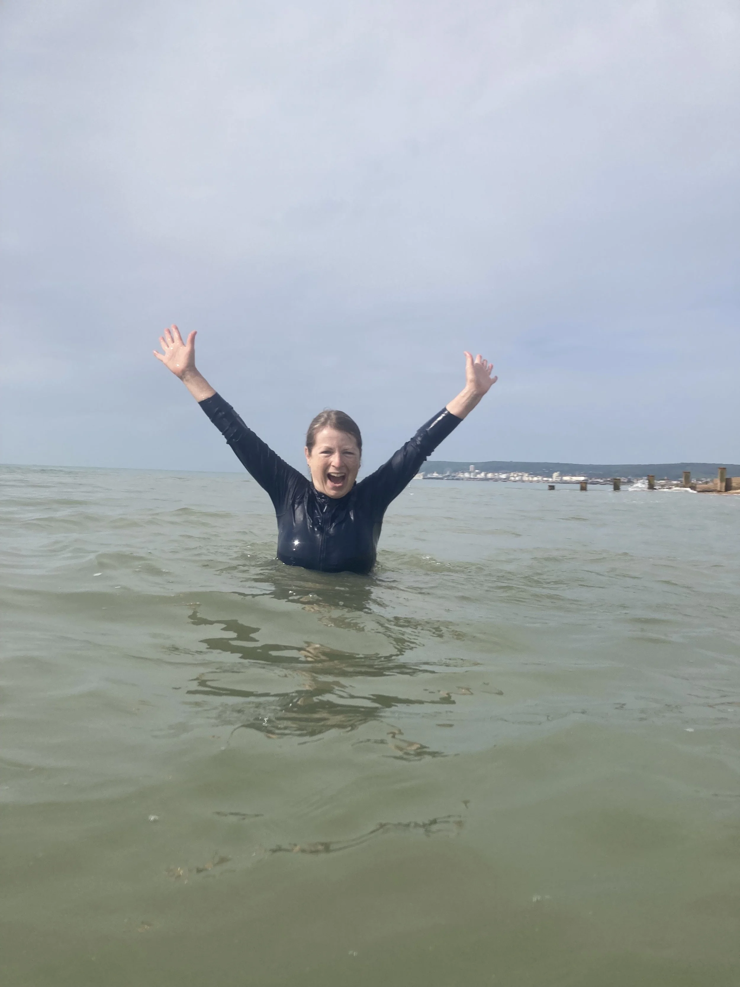 A woman Stands chest deep in the sea with her arms abover her head in a gesture of joy  