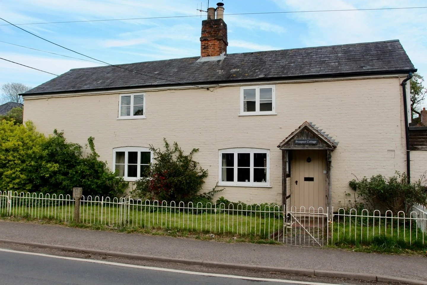 A two-story house with white brick walls, three white-framed windows on the second floor, two on the first, a small front garden with bushes, a white picket fence, and a wooden front door with a small porch and a sign that reads "Prospect Cottage".