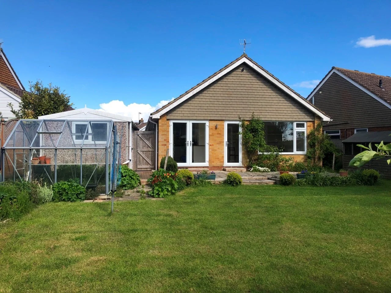 Rear view of a suburban house with a garden, featuring a greenhouse, plants, and a patio area under a blue sky.