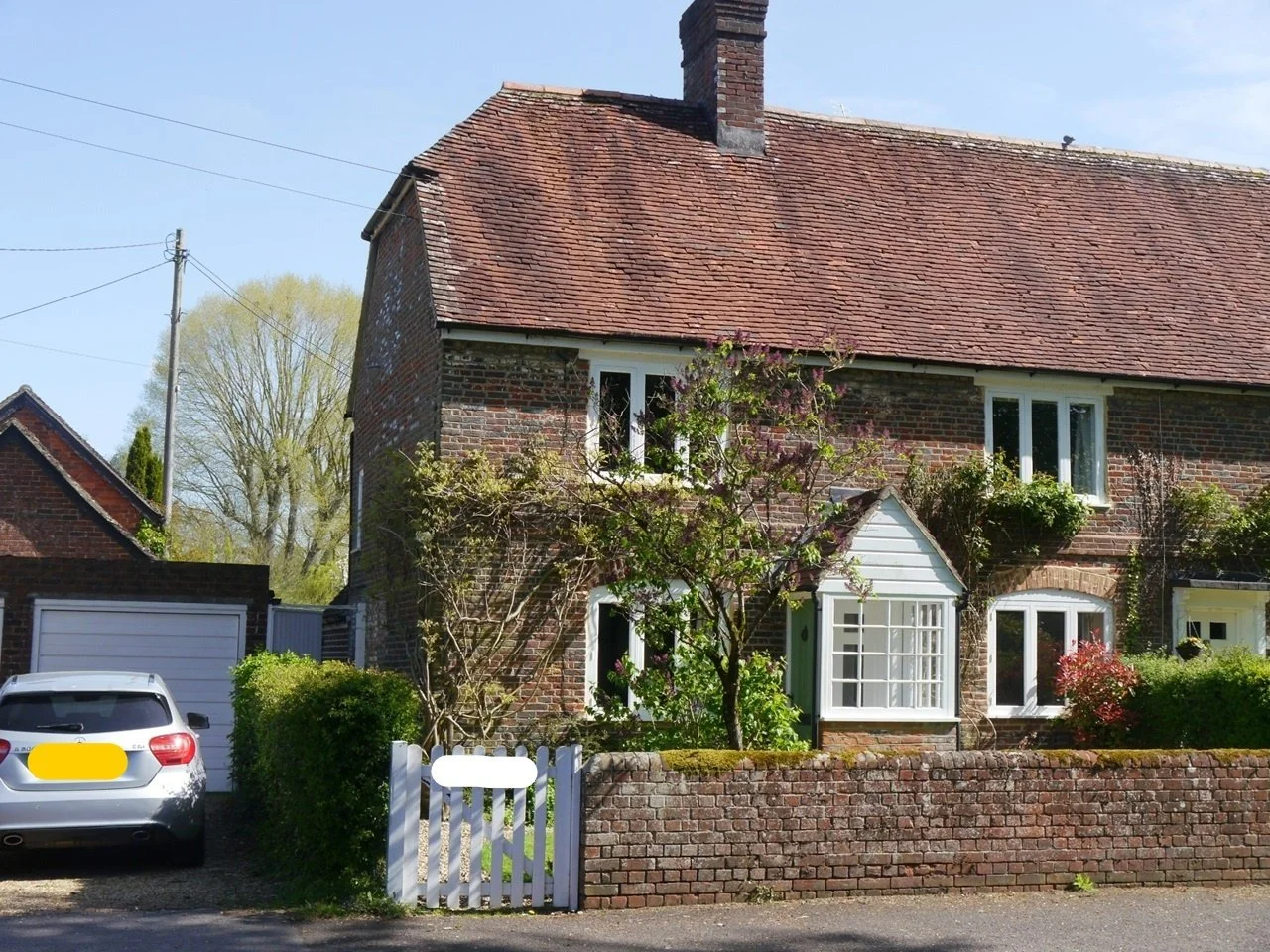 A brick house with a red tile roof and white-framed windows, surrounded by green bushes and trees, with a white picket fence and a silver car parked outside.