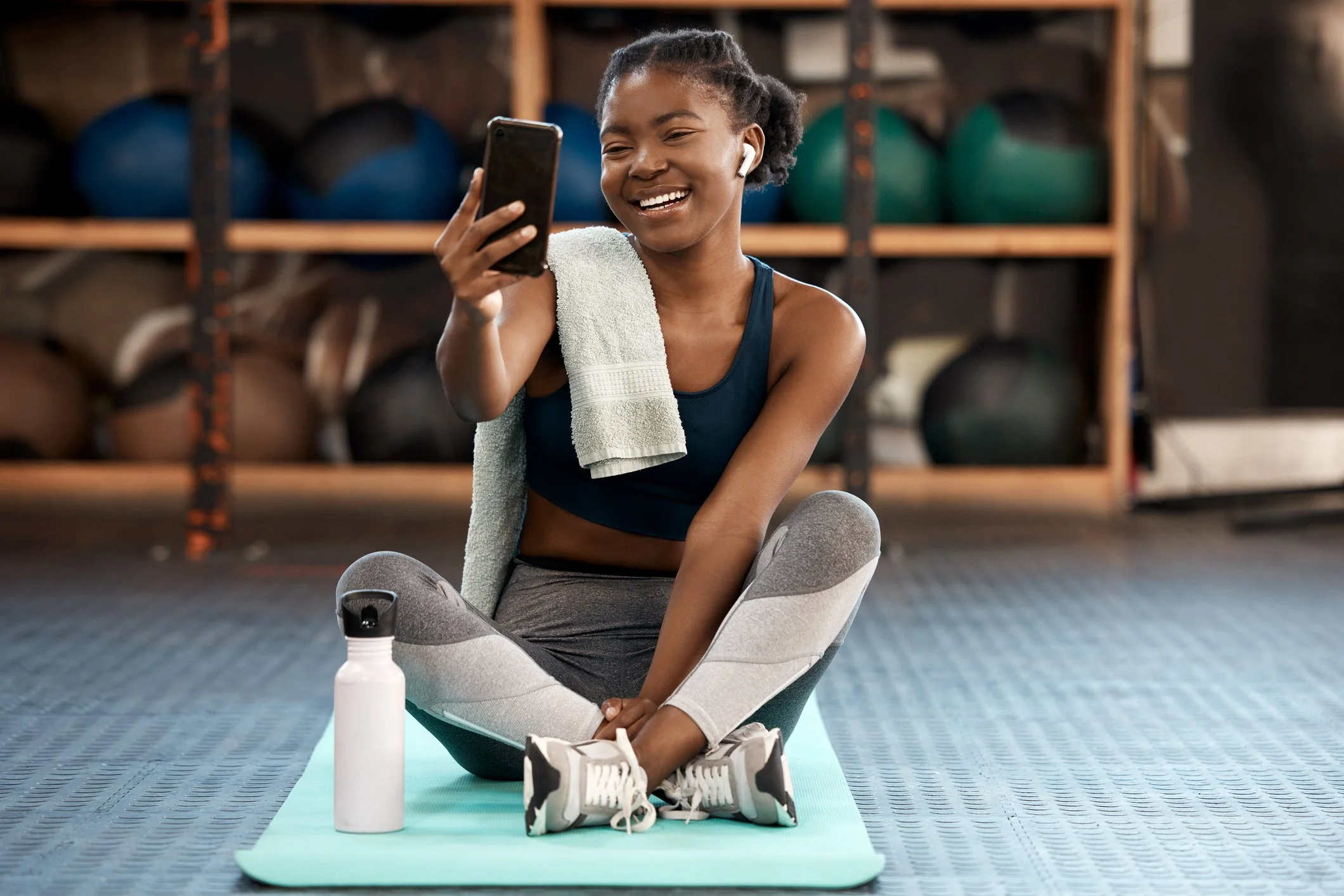 A teen sitting cross-legged on a yoga mat in a gym, smiling and taking a selfie with her smartphone. She has a towel over her shoulder, a water bottle in front of her, and is wearing workout clothes and wireless earbuds.