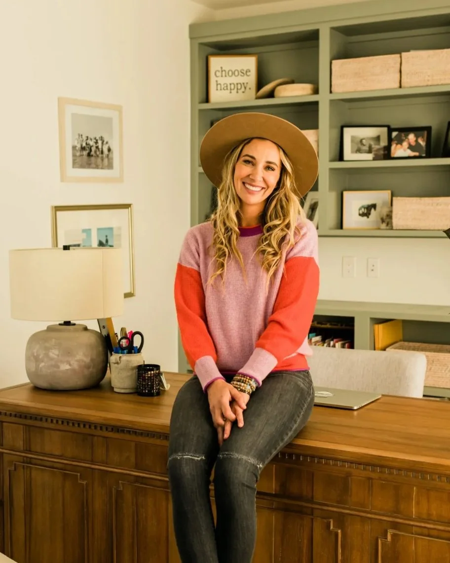 Co-founder with long blonde hair wearing a wide-brimmed hat, a colorful sweater, and ripped jeans, sitting on a wooden desk in a home office with bookshelf and framed photos behind her.
