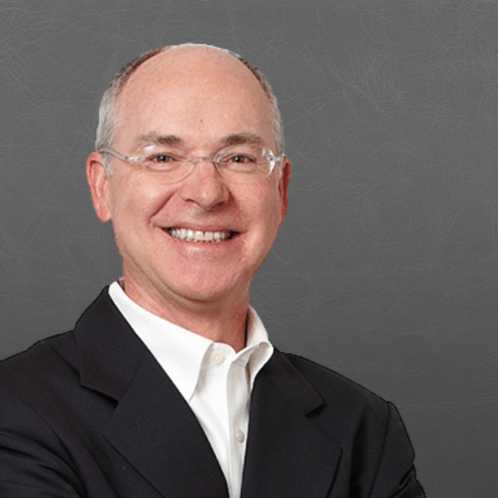 A smiling man with glasses, dressed in a black suit and white shirt, standing against a plain gray background.