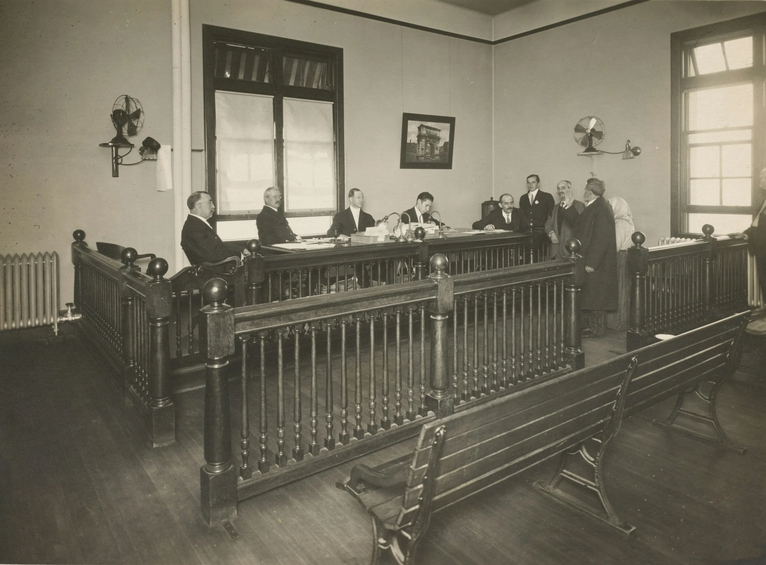 A black-and-white photograph of a courtroom with several men sitting and standing around a wooden railing, with large windows and vintage-style fan and lighting fixtures on the walls.
