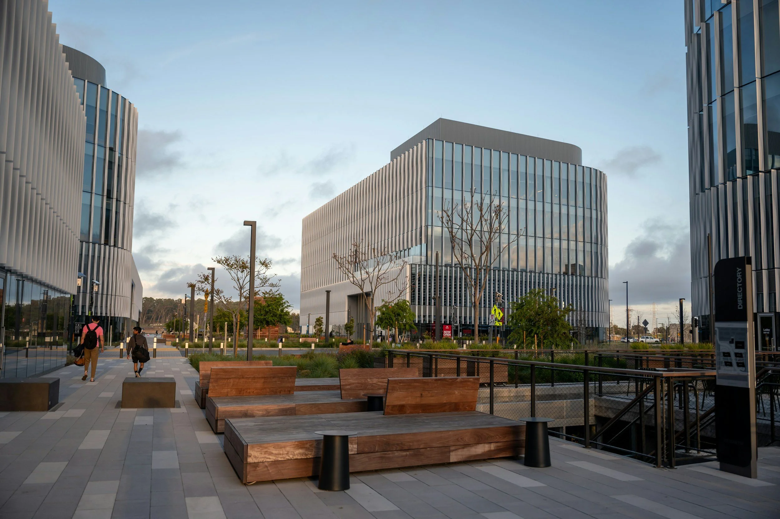 Modern office buildings with glass facades and an outdoor paved plaza with benches, trees, and people walking.