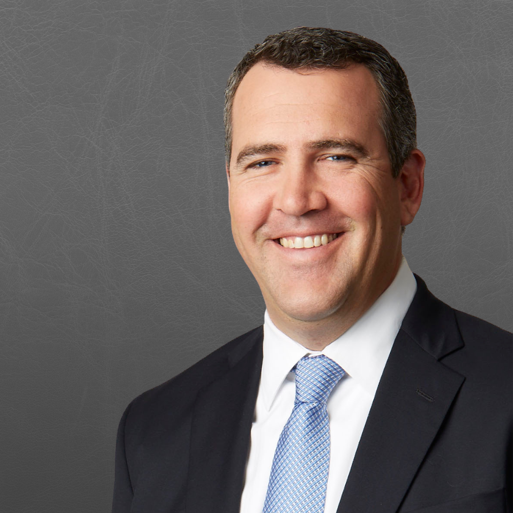 A smiling man dressed in a black suit, white shirt, and light blue tie against a gray textured background.