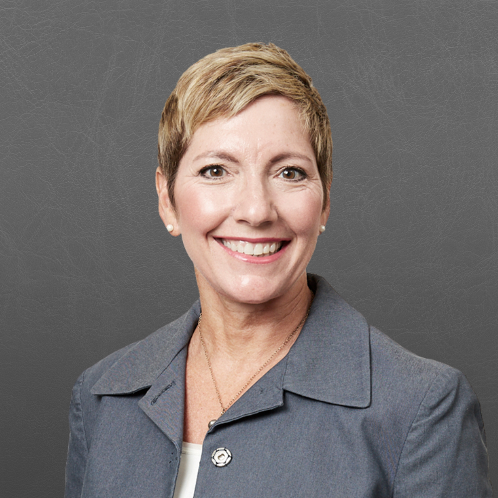 A smiling woman with short blonde hair, wearing a gray blazer, earrings, and a necklace, against a gray background.