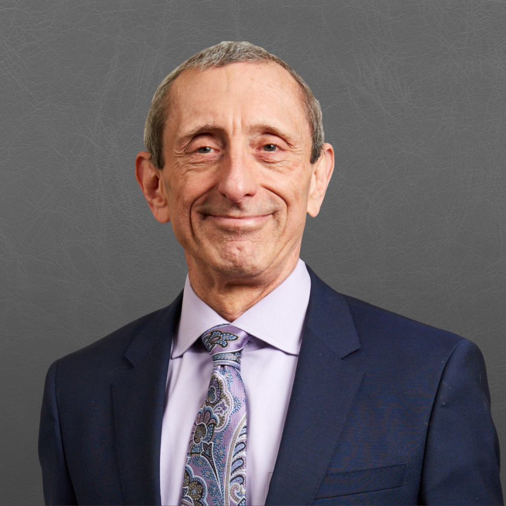 A smiling older man in a dark suit, lavender shirt, and a patterned tie against a gray textured background.