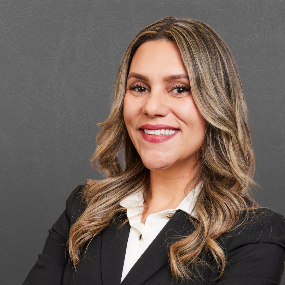 A professional woman with long, wavy blonde hair, wearing a black blazer over a white shirt, smiling against a gray textured background.