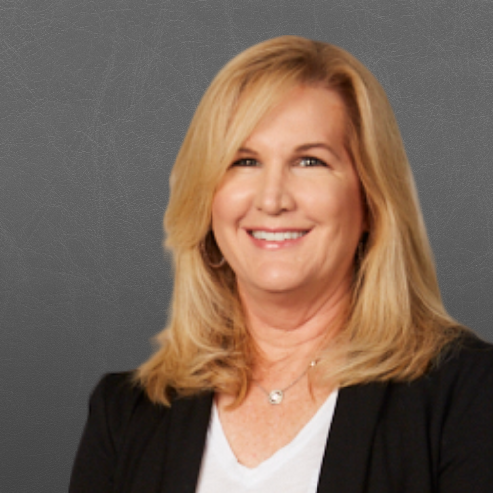 Portrait of a smiling woman with blonde hair, wearing a black blazer and a white top, set against a gray background.