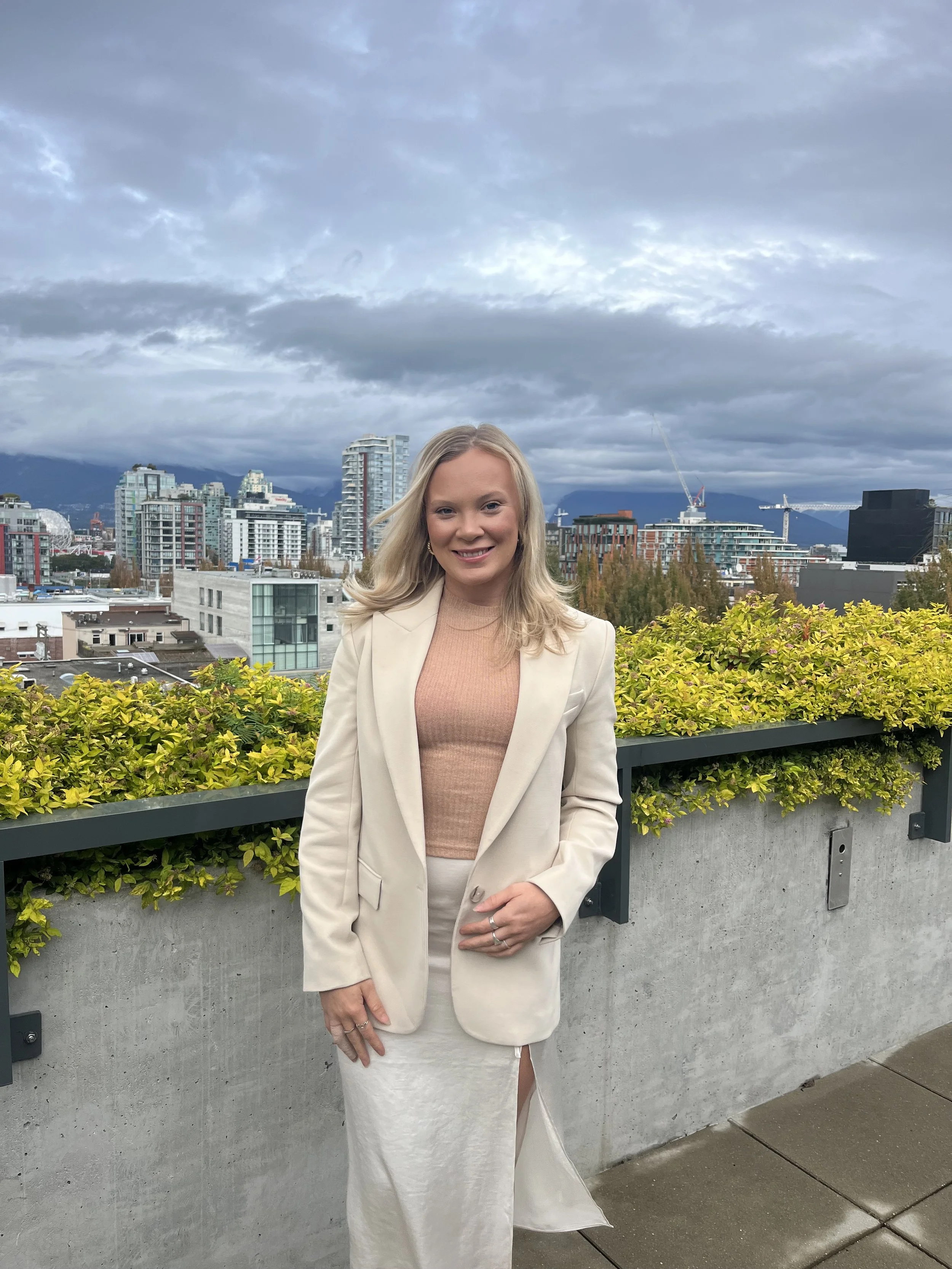 A woman standing on a rooftop with city buildings and mountains in the background, wearing a beige blazer and skirt with a pink top, under a cloudy sky.