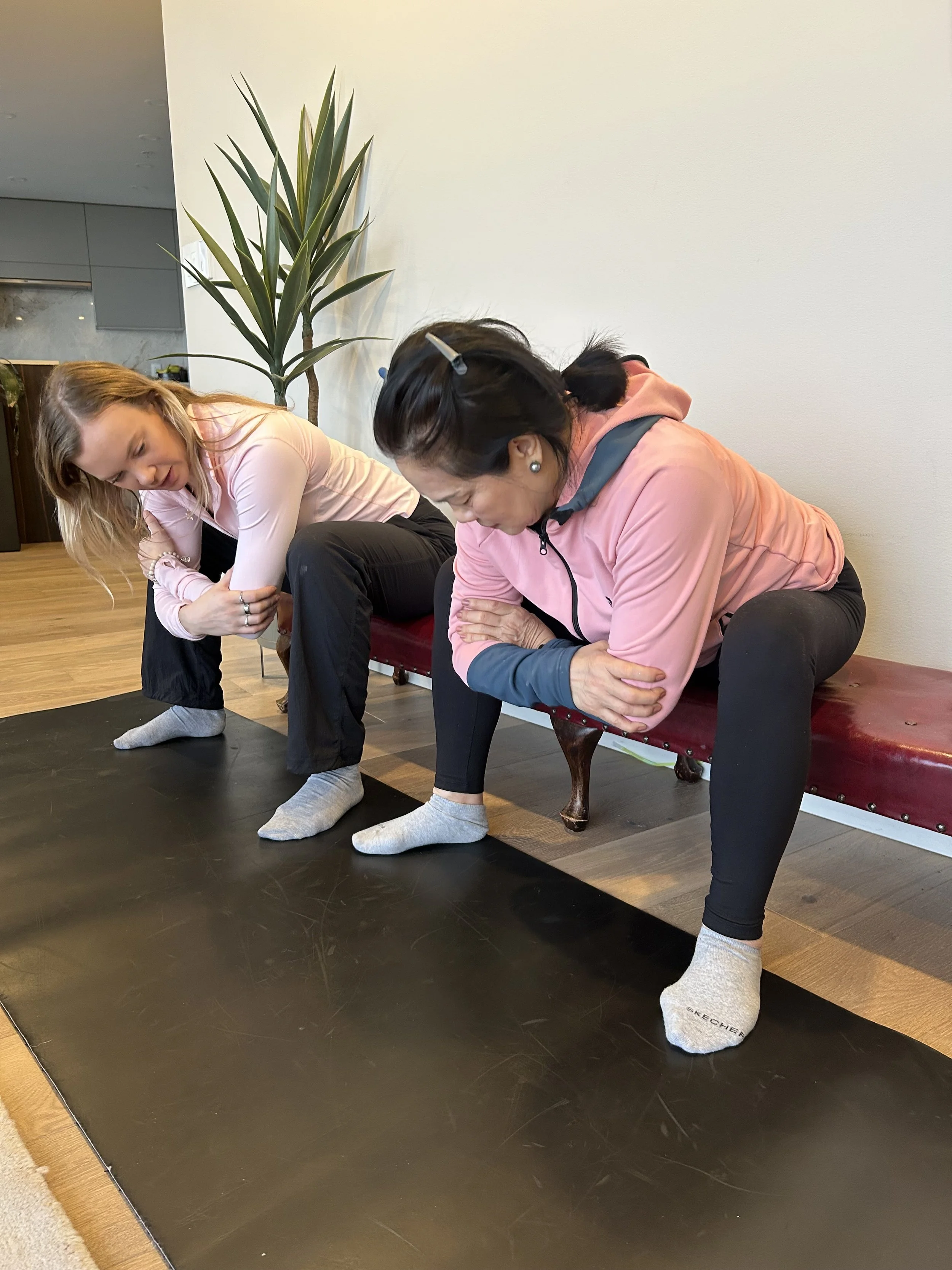 Two women sitting on a bench, stretching their legs and leaning forward, with black exercise mats on the floor in front of them.