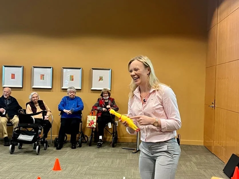 A woman with blonde hair, wearing a pink shirt and gray pants, smiles and holds a yellow stuffed animal, standing in a room with four elderly people seated in the background, with framed artwork on the wall.