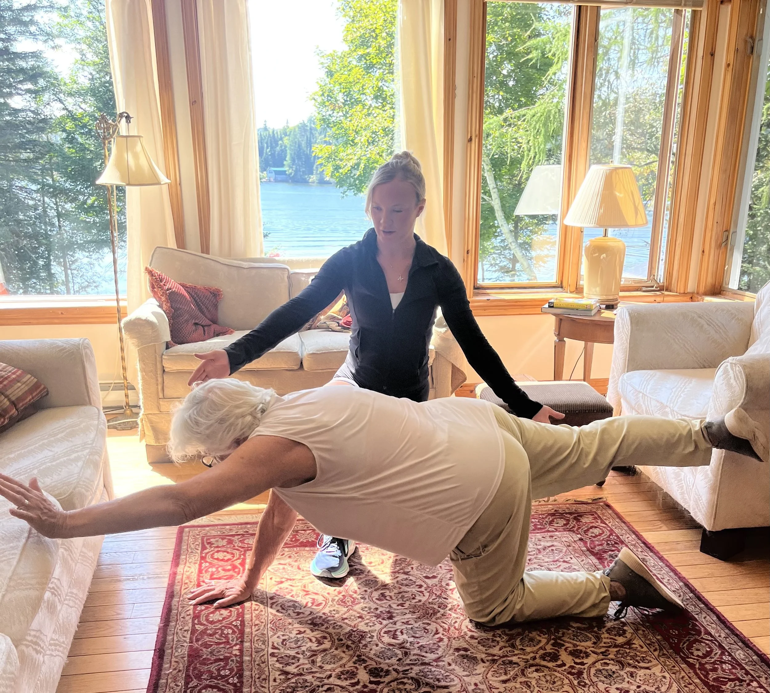 A woman giving an elderly woman a fitness or yoga pose in a bright living room with large windows, a view of a lake, and cozy furniture.