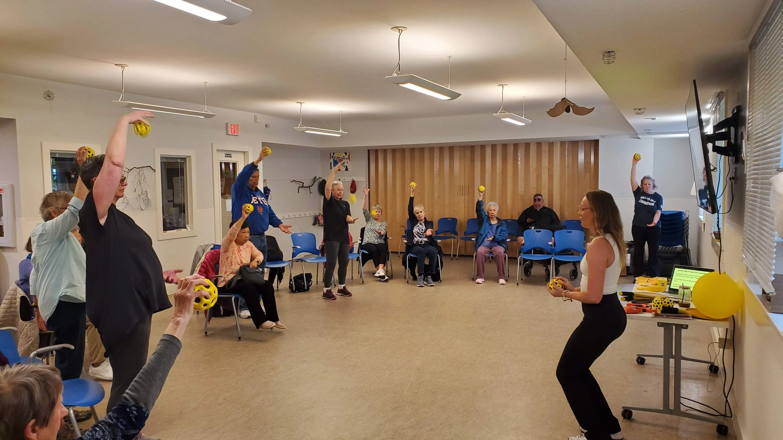 A woman in black pants and a black shirt is giving a presentation to a group of elderly people seated in chairs in a bright room. The presentation slide on the screen displays a greeting to residents, logos, and an image of a woman in a graduation cap.