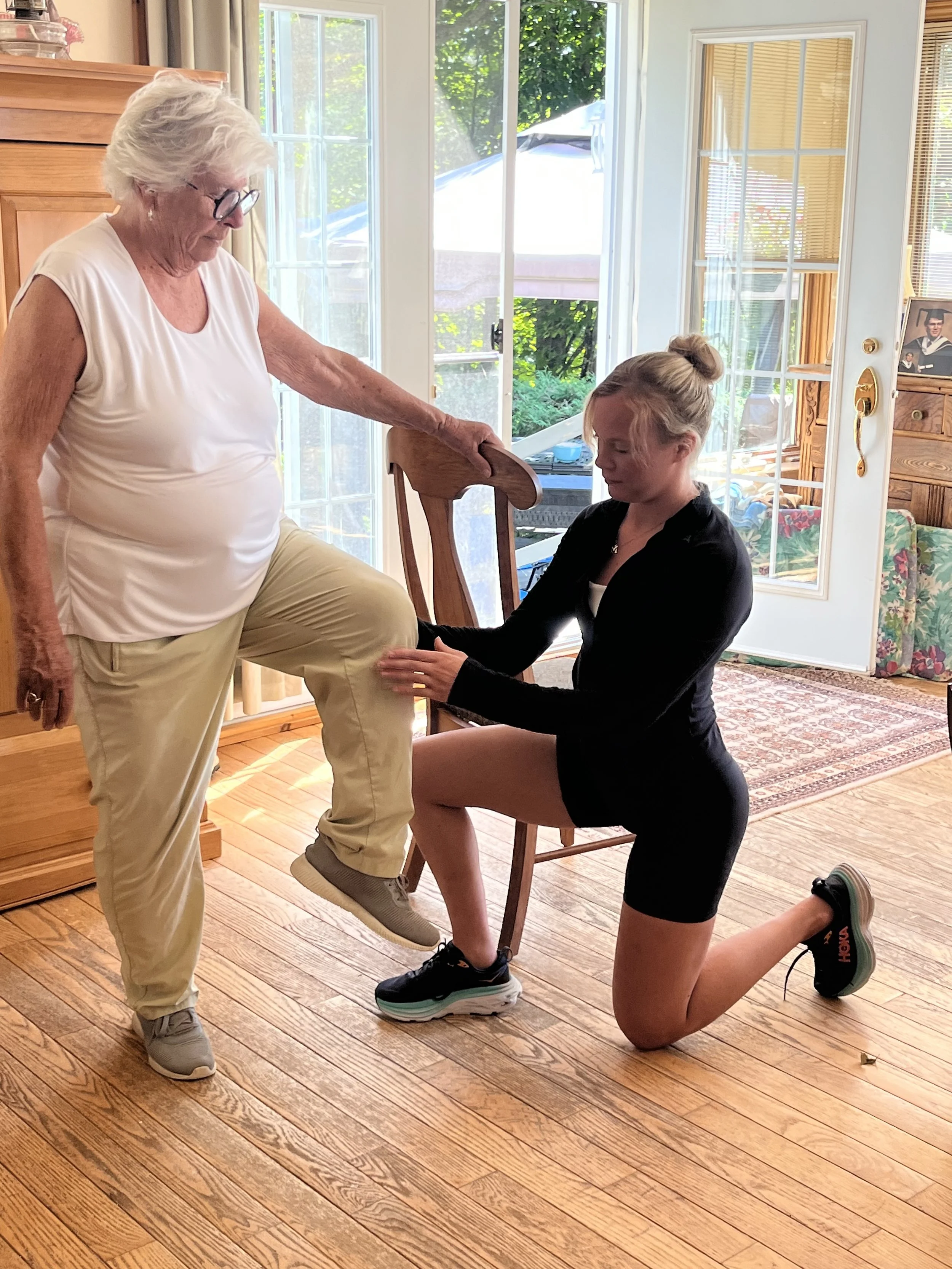 A young woman in black workout clothes kneels on one knee while holding a chair for support, and an older woman stands nearby with her foot resting on the young woman's knee, indoors near glass doors with a view of greenery outside.