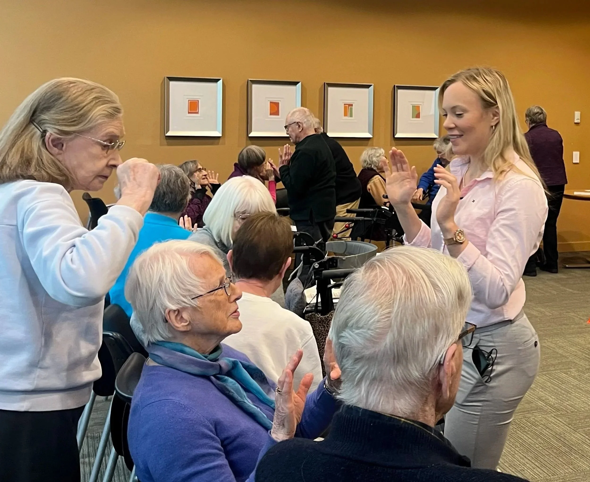 Group of elderly people and caregivers engaged in a therapeutic or social activity in a community room, with some participants raising their hands and others sitting and listening, decorated with abstract artwork on the wall.
