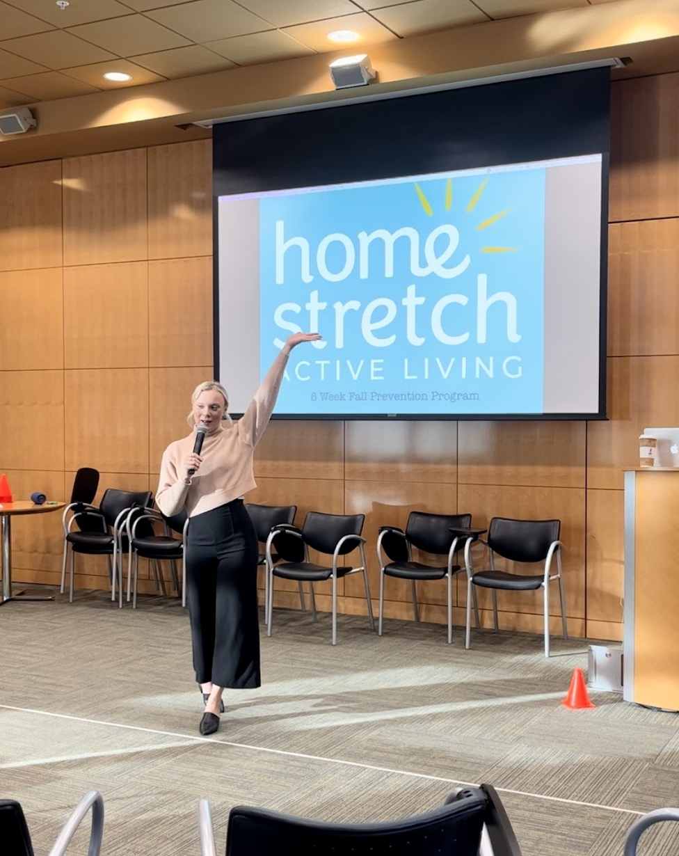 A woman standing in front of a large screen in a conference room, giving a presentation about a program called 'Home Stretch Active Living'. She is holding a microphone and gesturing with her right hand, with several chairs behind her and a few orange cones on the floor.