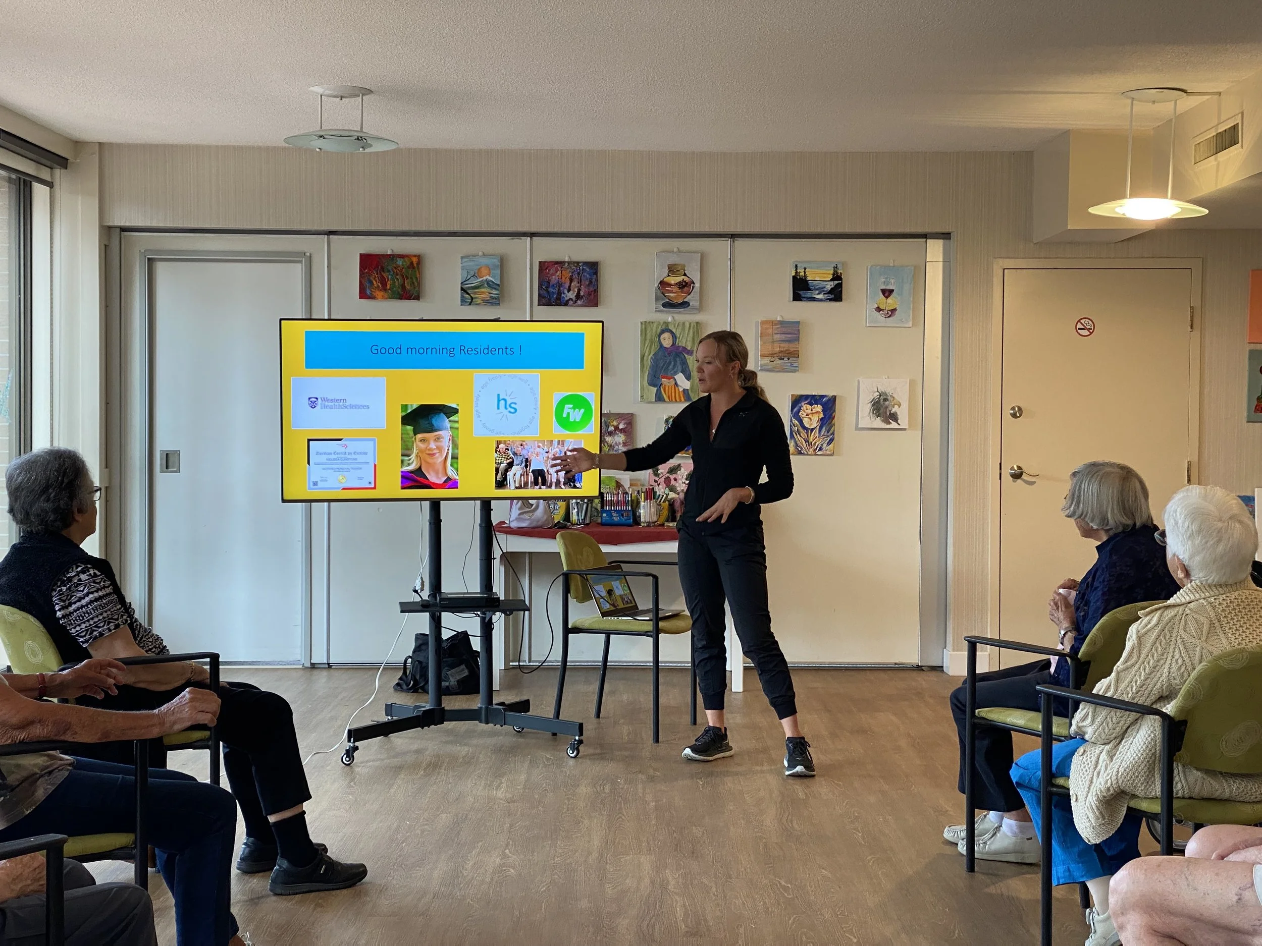 A woman in black pants and a black shirt is giving a presentation to a group of elderly people seated in chairs in a bright room. The presentation slide on the screen displays a greeting to residents, logos, and an image of a woman in a graduation cap.