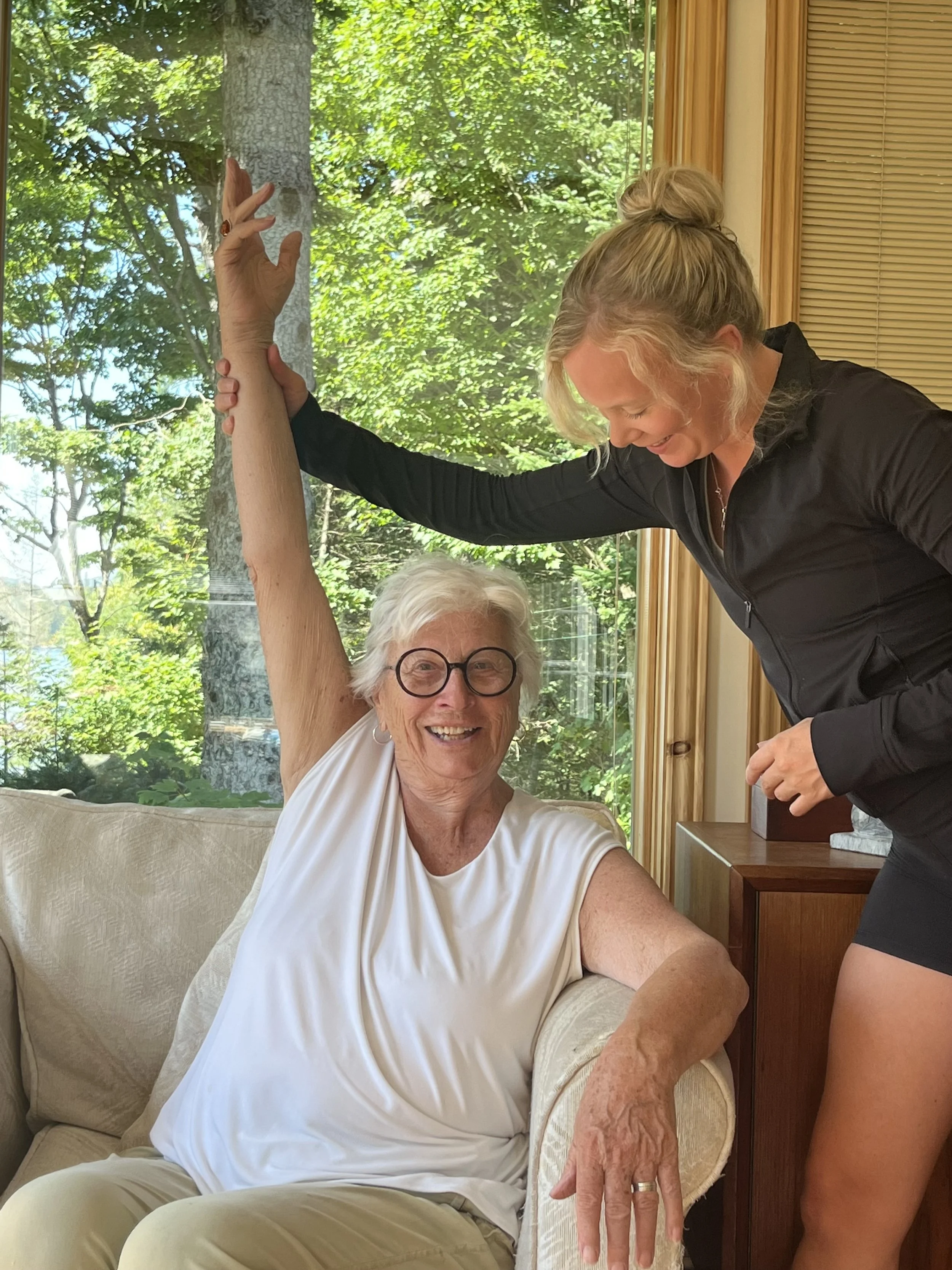 An elderly woman with white hair, glasses, and a sleeveless white top sitting on a beige couch with a smile, while a younger woman stands beside her, holding her raised arm. The setting appears to be a bright room with large windows revealing green trees outside.