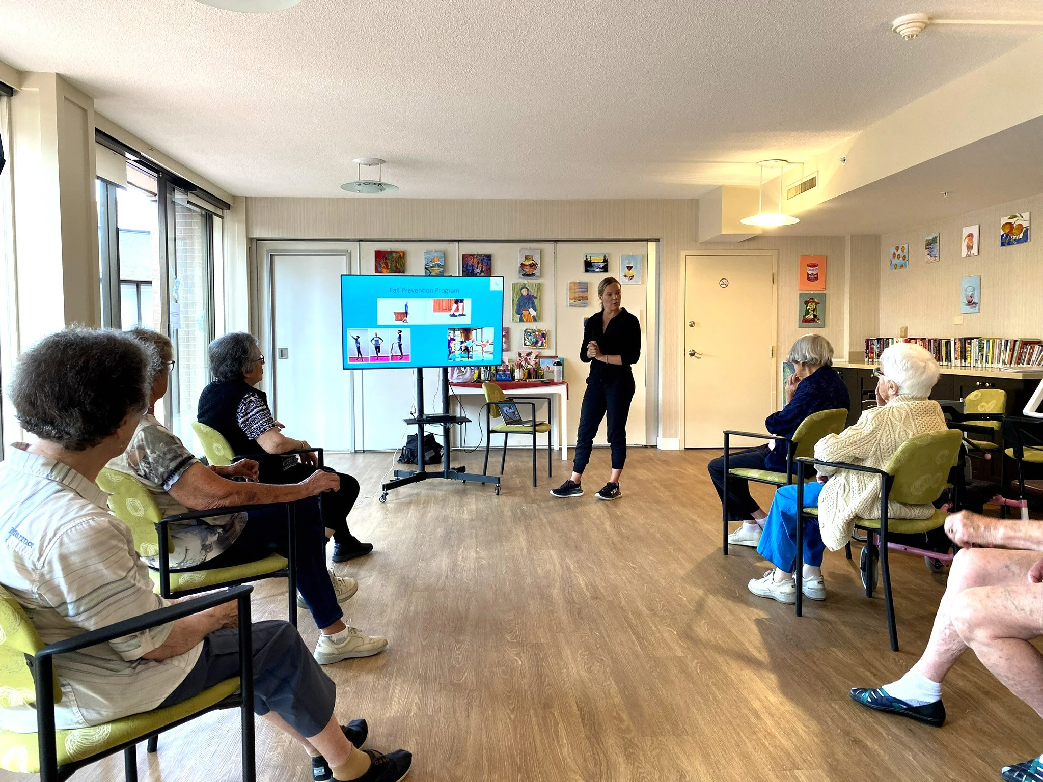 A group of elderly people sitting in chairs watching a fitness instructor give a presentation on fall prevention in a common room with colorful artwork on the walls