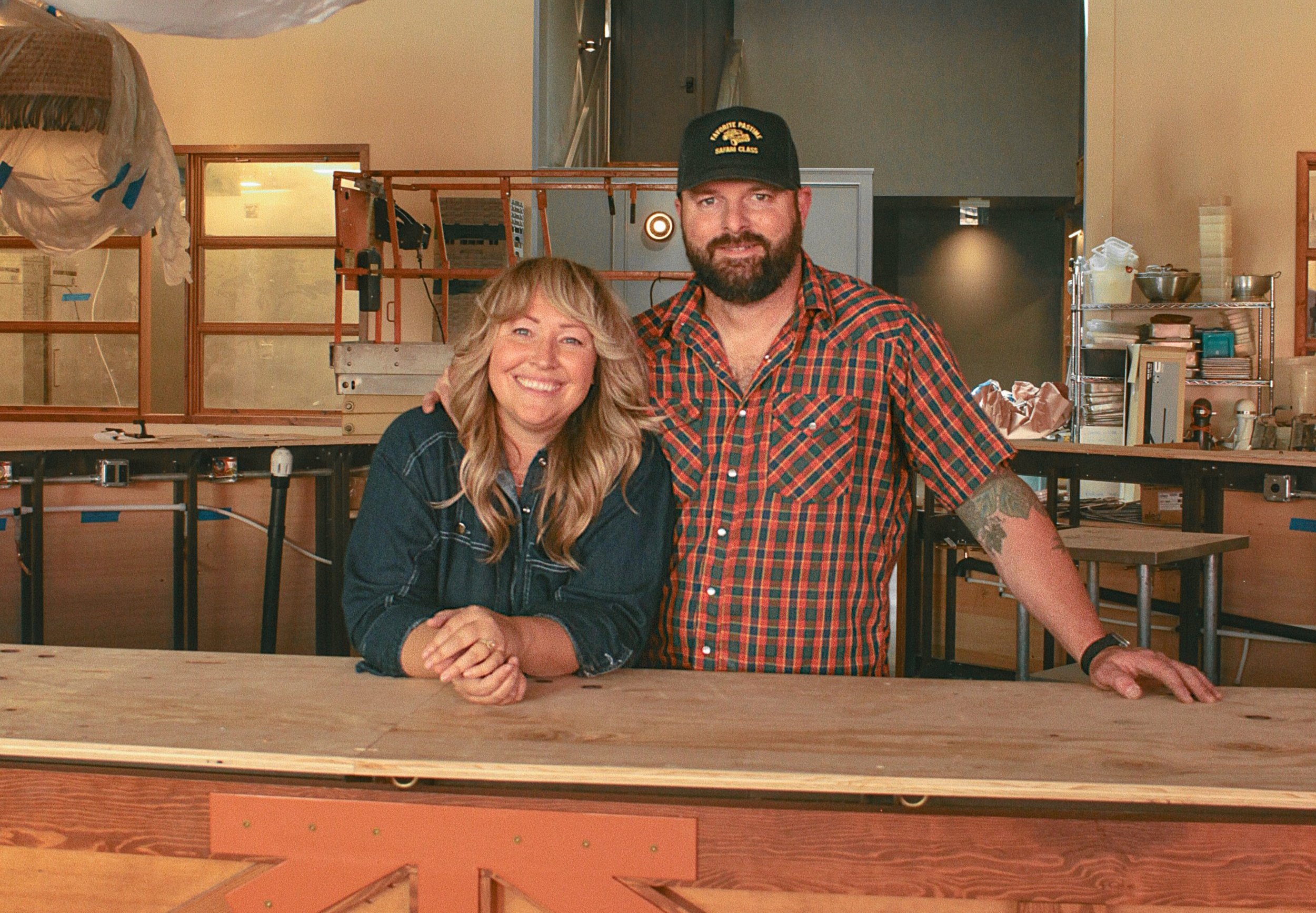 A woman and a man standing at a wooden counter in a kitchen or workshop, smiling at the camera. The woman has long blonde hair and is wearing a dark denim shirt, while the man has a beard, tattoos on his arm, and is wearing a red and green plaid shirt and a black baseball cap.