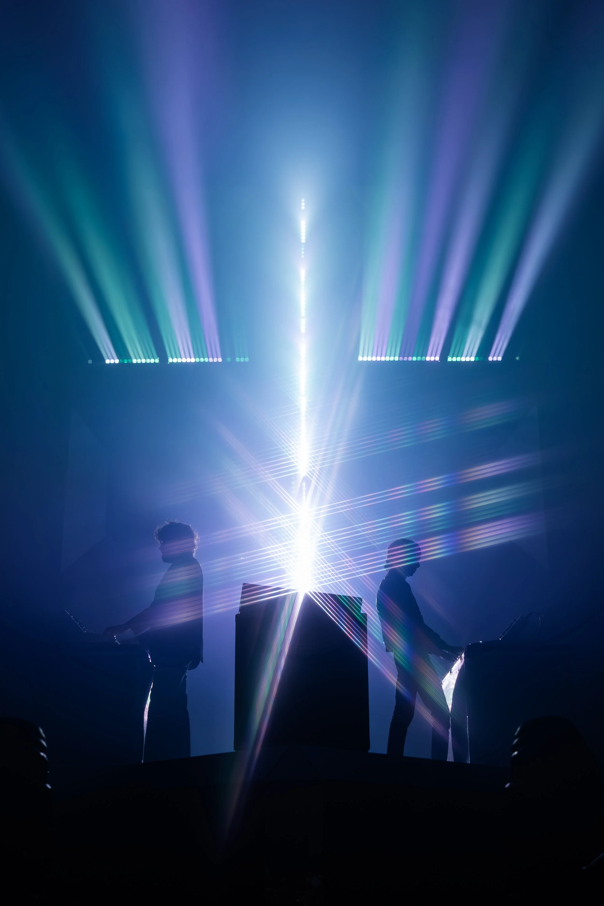 Deux DJ avec des équipements musicaux dans une scène de spectacle avec des jeux de lumières colorés et des faisceaux lumineux brillante au centre.