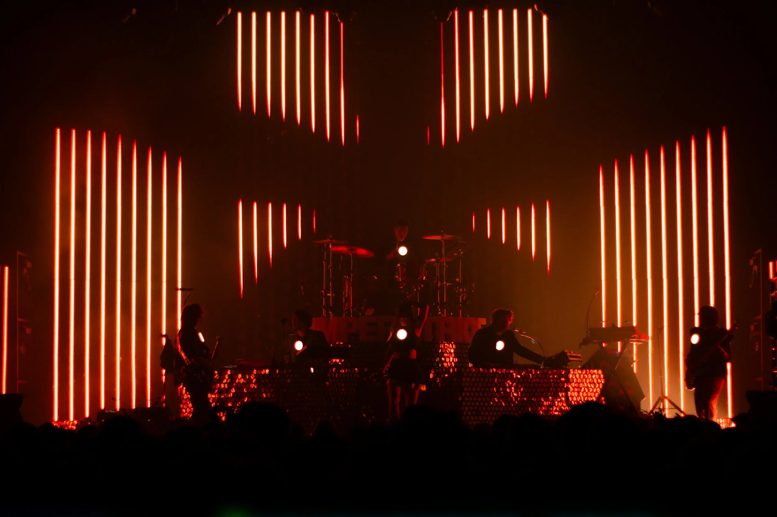 Performance musicale en direct sur scène avec lumières rouges verticales et silhouettes de musiciens.