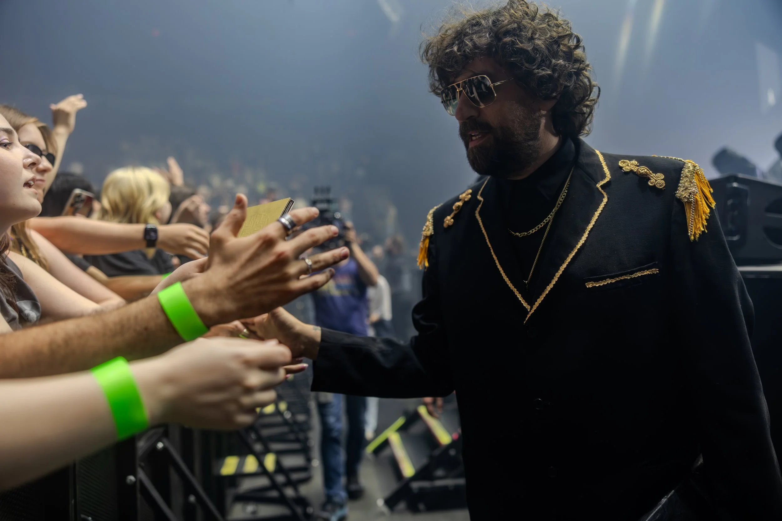 Un homme avec des cheveux bouclés, portant des lunettes de soleil et une veste noire ornée de broderies dorées, serre la main de plusieurs personnes lors d'un concert ou d'un événement public.