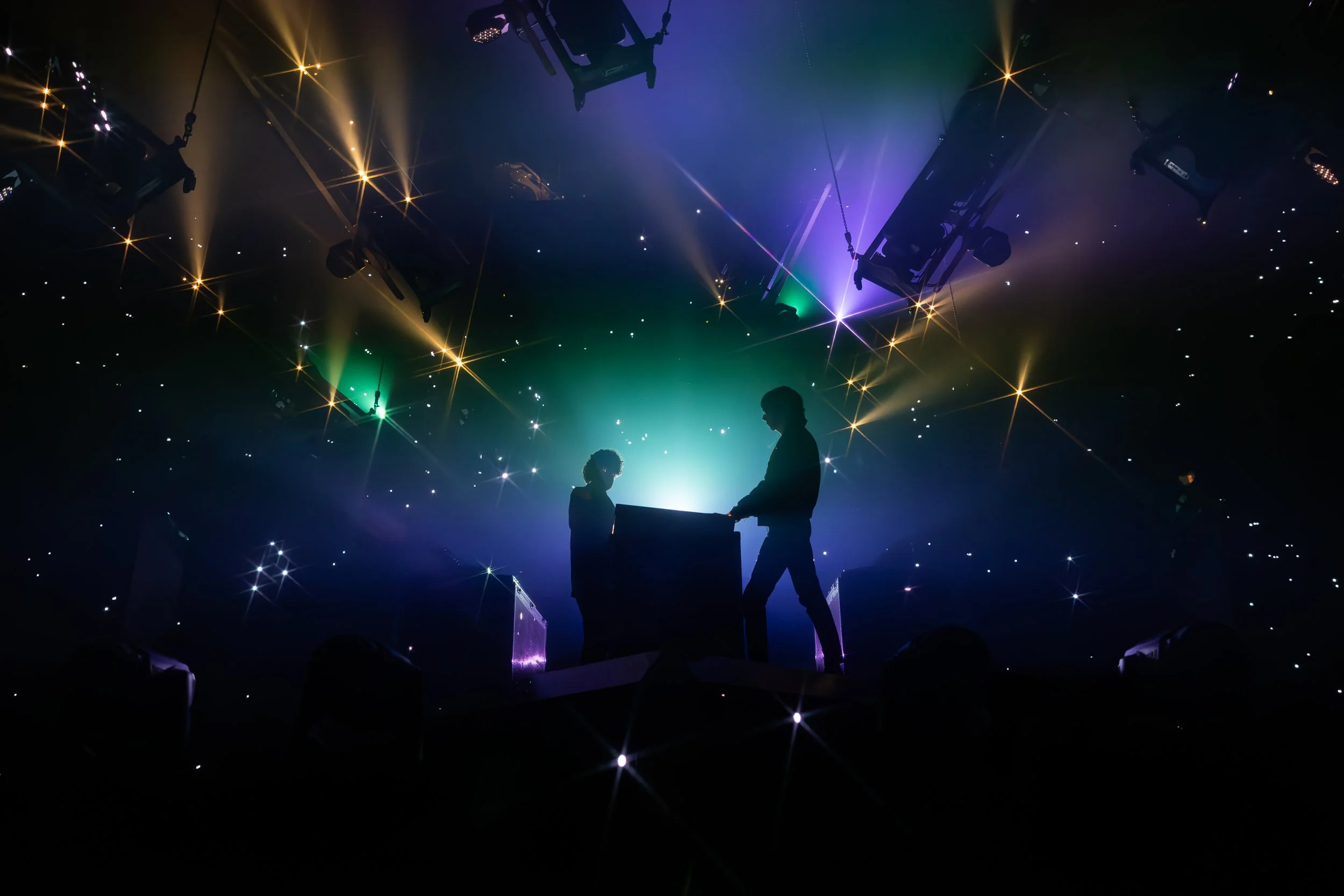 Deux personnes en silhouette derrière un DJ, éclairage coloré et effets lumineux sur scène lors d'un concert ou spectacle de musique.