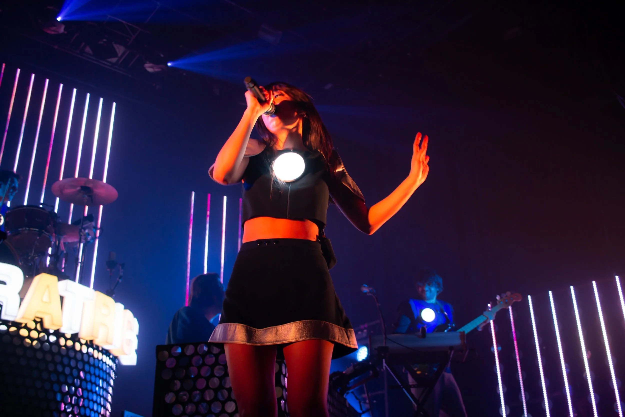 Jeune femme chantant au microphone sur scène, avec un groupe musical en arrière-plan, éclairage coloré. 