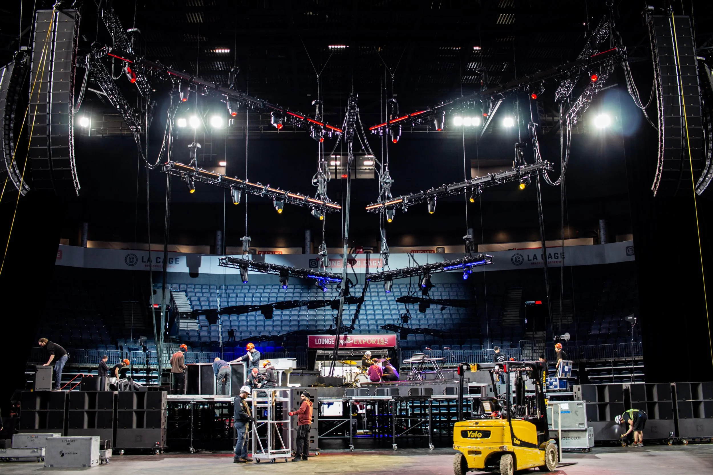 Préparation d'une scène de spectacle avec des travailleurs installant du matériel, un éclairage sophistiqué suspendu au plafond, et des sièges en arrière-plan dans une salle de concert ou un théâtre.