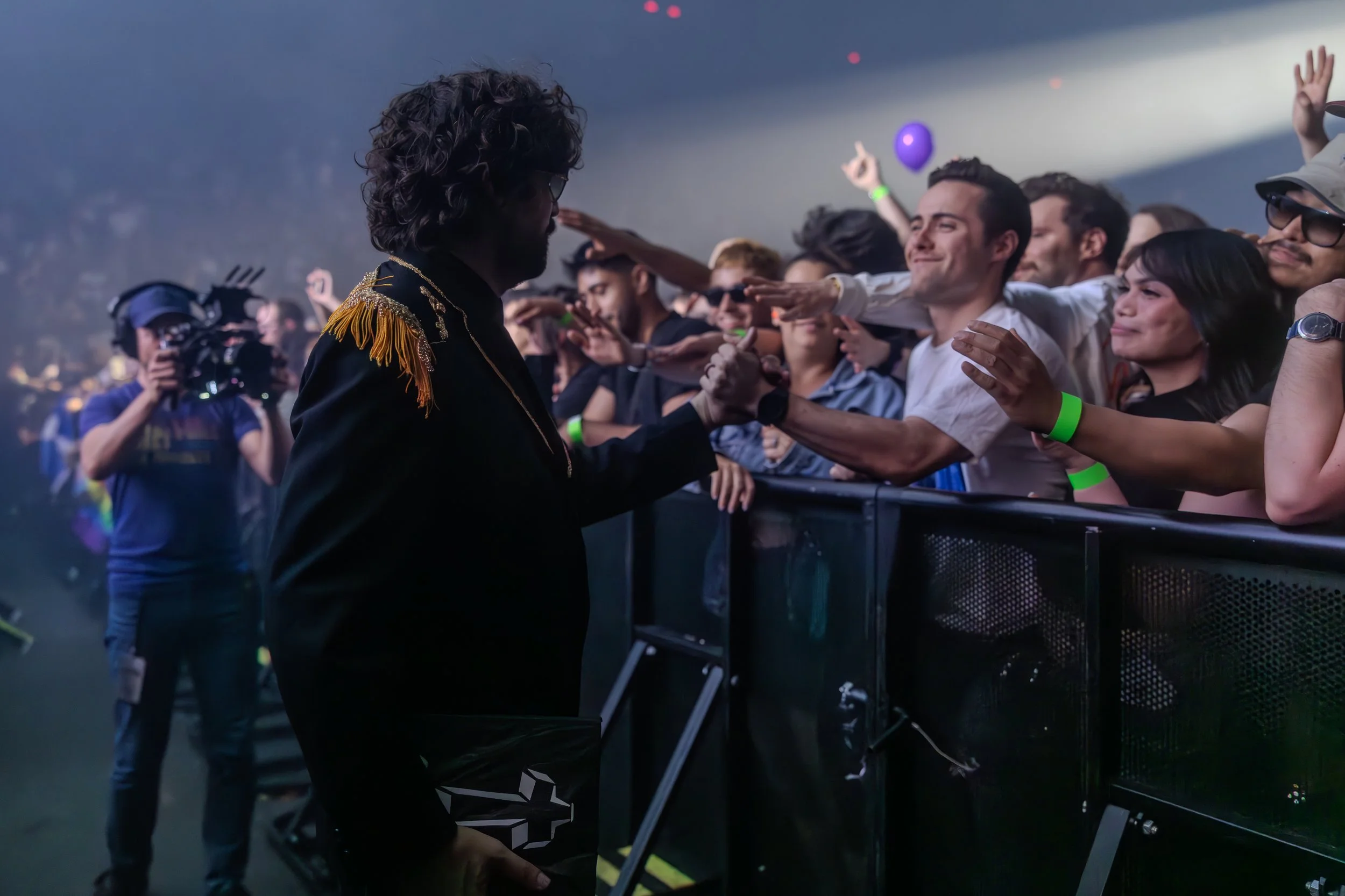 Un homme avec des cheveux bouclés et portent une veste noire avec des épaulettes dorées, regarde un groupe de personnes au public lors d'un concert ou un événement en intérieur, avec des gens qui tendent la main vers lui, certains portant des bracele