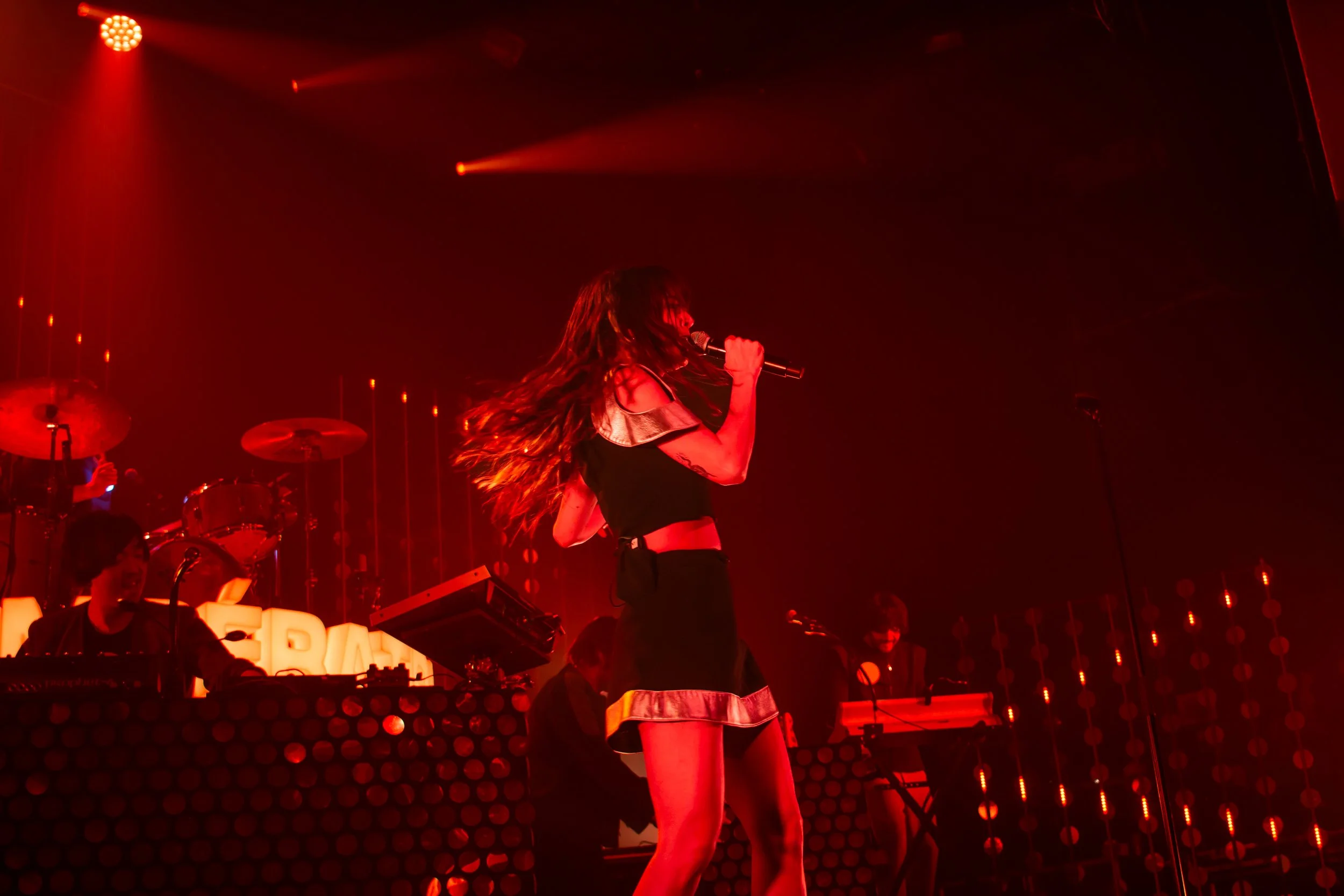 Une chanteuse dans une scène de concert, avec des musiciens en arrière-plan, tous sous un éclairage rouge intense.
