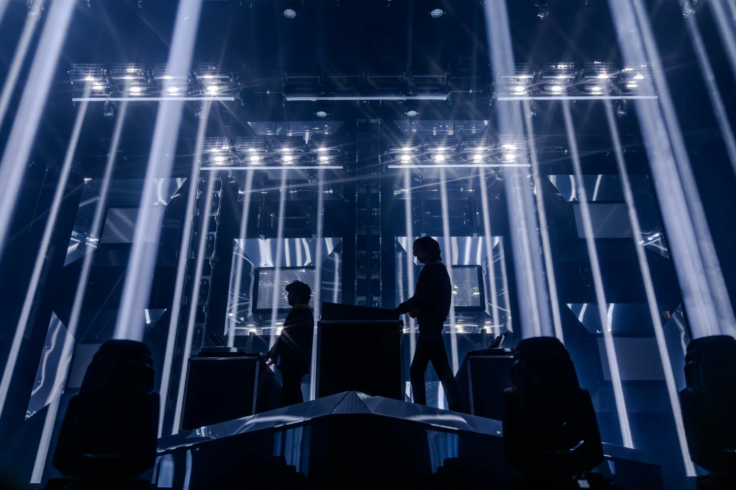 Silhouettes de deux DJ ou techniciens en train de préparer ou gérer la scène dans une salle de concert ou de spectacle futuriste, avec un système d'éclairage complexe et des écrans LED derrière eux, créant une atmosphère high-tech et moderne.