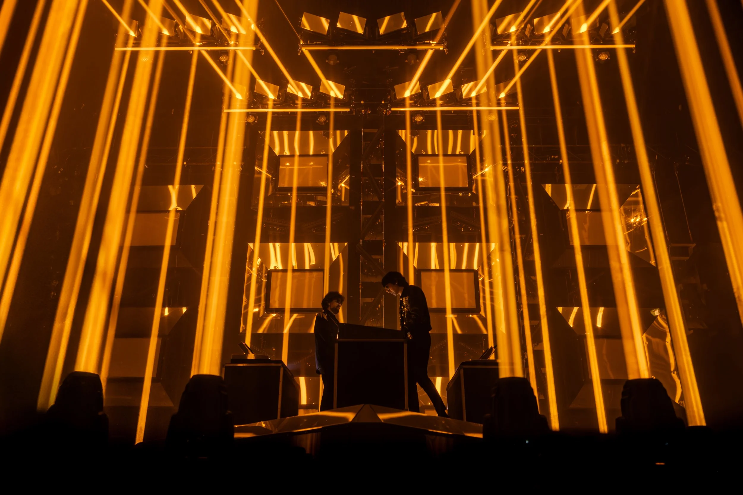 Deux personnes sur scène entourées de lampes jaunes et de cadres lumineux dans un environnement sombre, ambiance de spectacle ou de concert.