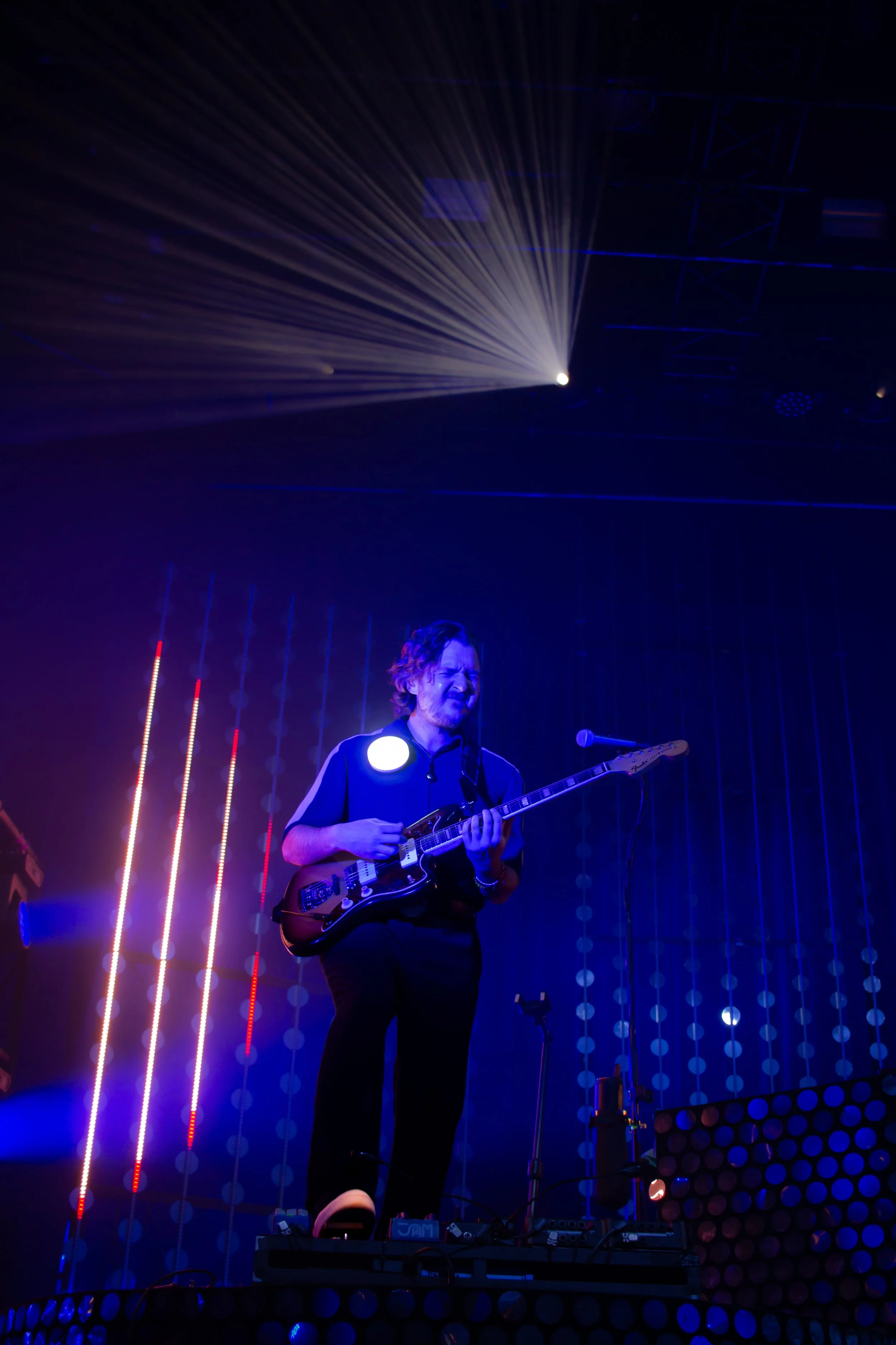 Un musicien joue de la guitare électrique sur une scène éclairée de lumières colorées et de néons, avec un fond lumineux et un faisceau lumineux intense derrière lui.