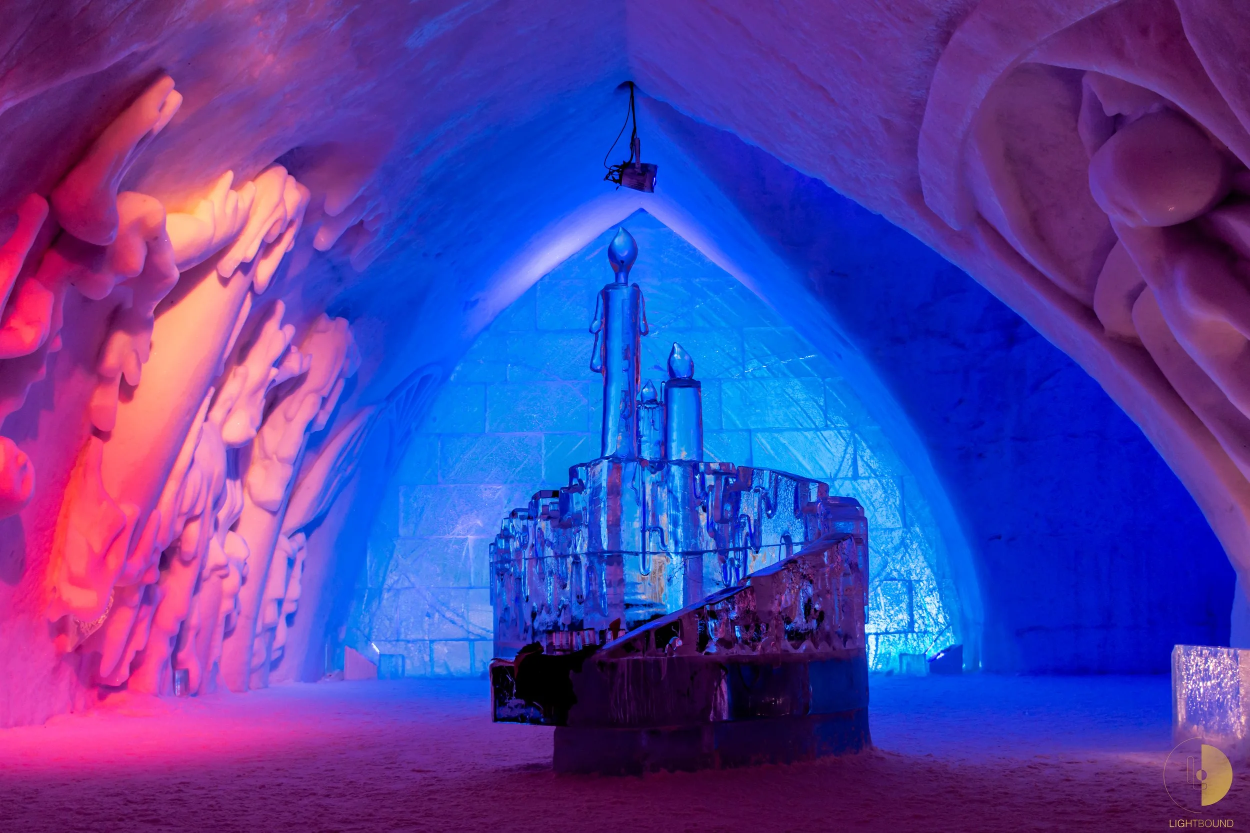 Une salle d'ascension glacée de l'ICEHOTEL, ornée de sculptures de glace illuminées par des lumières colorées, avec une structure centrale en glace.
