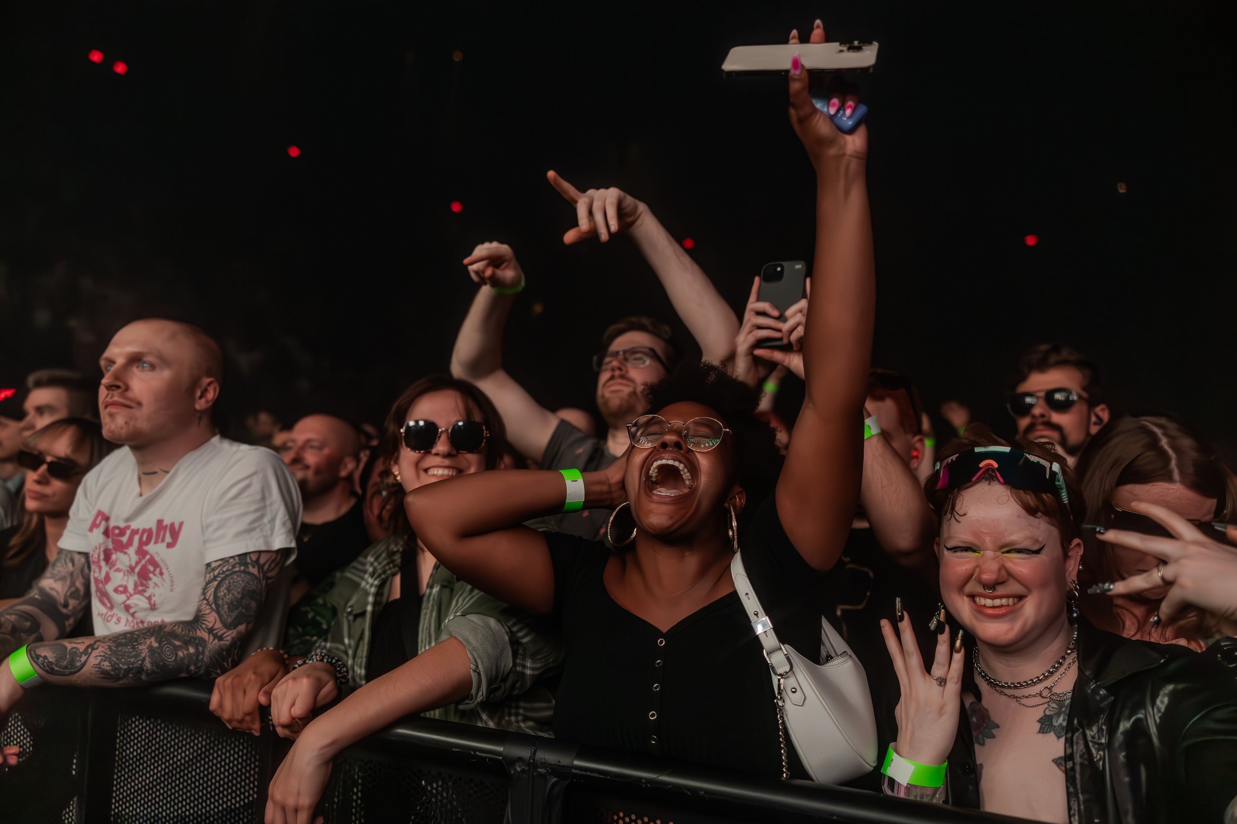 Groupe de personnes riant et chantant lors d'un concert, levant les mains et portant des accessoires comme des lunettes de soleil et des écouteurs, dans une ambiance festive.