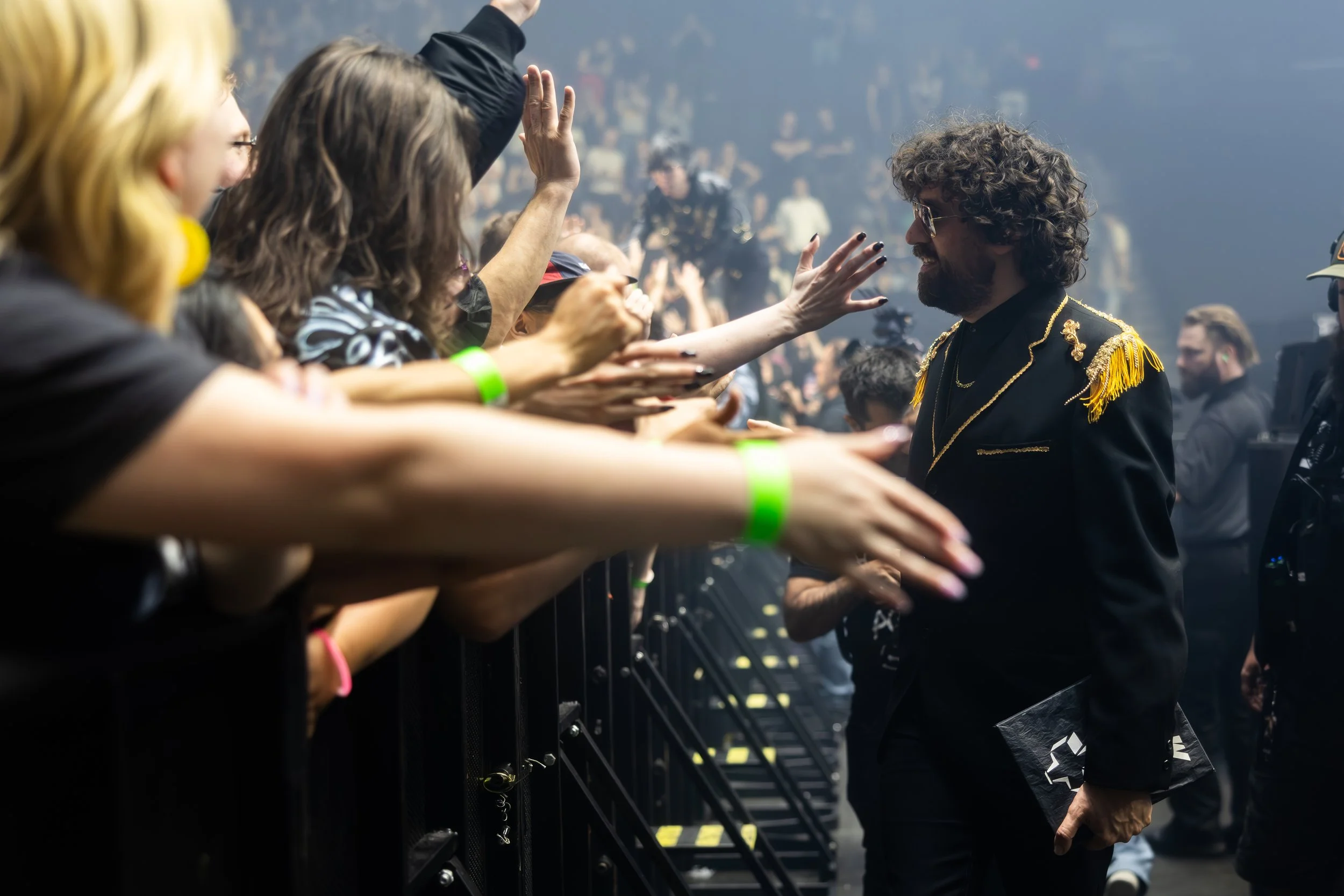 Un homme avec des cheveux bouclés, portant une veste noire avec des épaulettes dorées, serre la main de plusieurs personnes du public lors d'un concert ou d'un événement, avec une scène en arrière-plan.