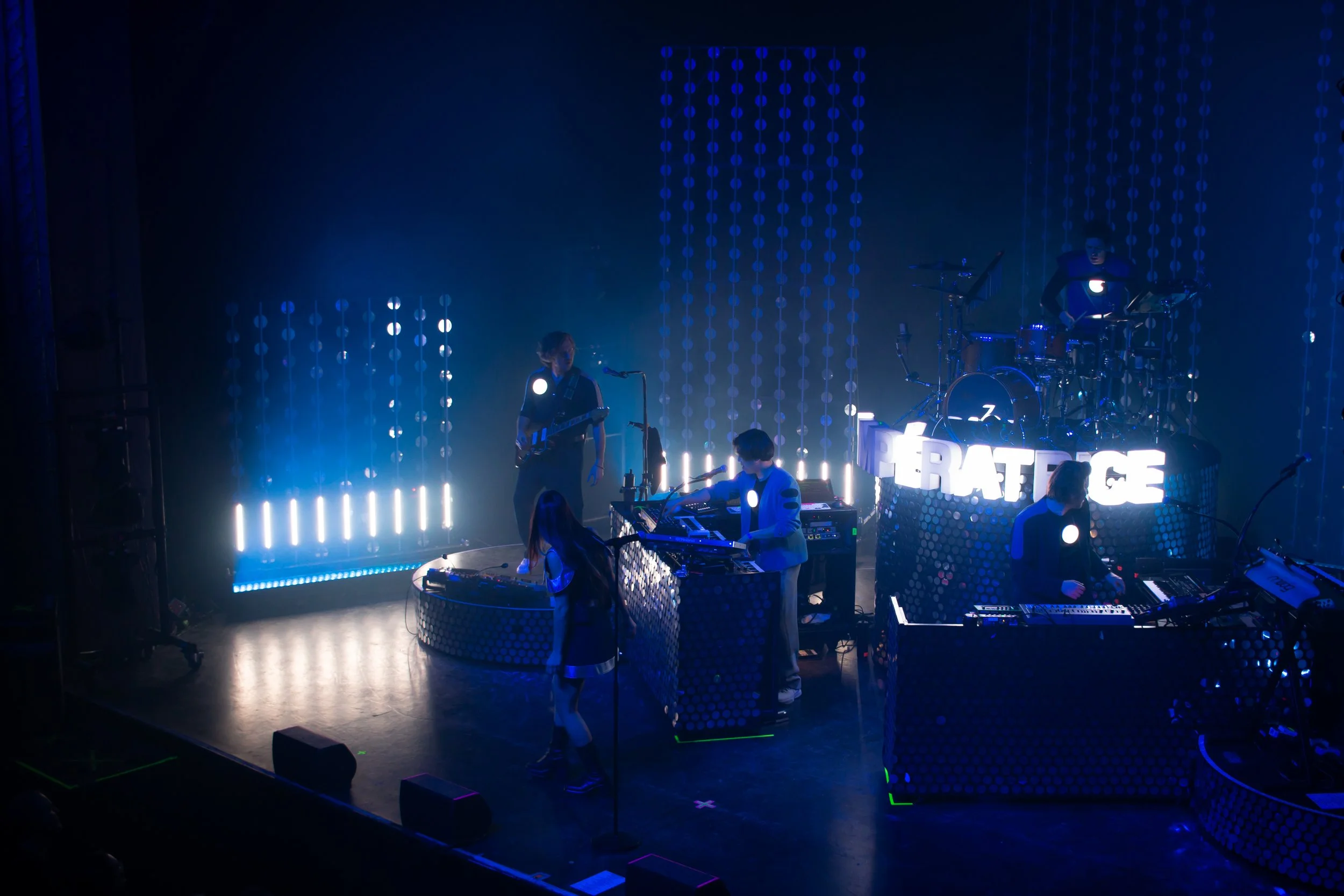 Un groupe de musiciens en concert, avec des membres jouant de la guitare, du clavier, de la batterie et un chanteur, sur scène illuminée de lumière bleue et blanche, avec un panneau lumineux affichant le mot "PERFORMANCE".