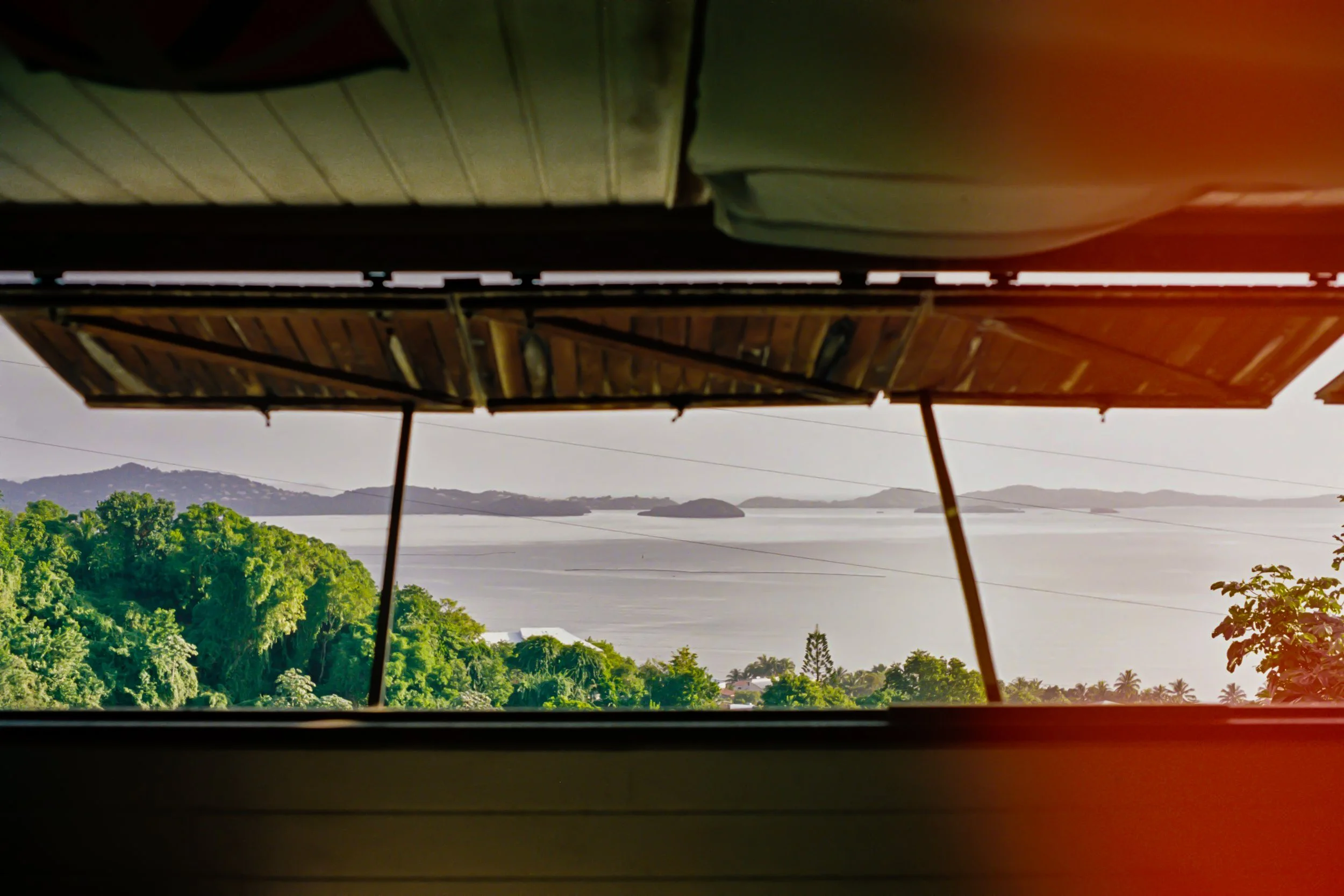 Vue depuis une fenêtre montrant de la verdure, la mer et des îles au loin, avec un toit en bois partiellement visible en haut.