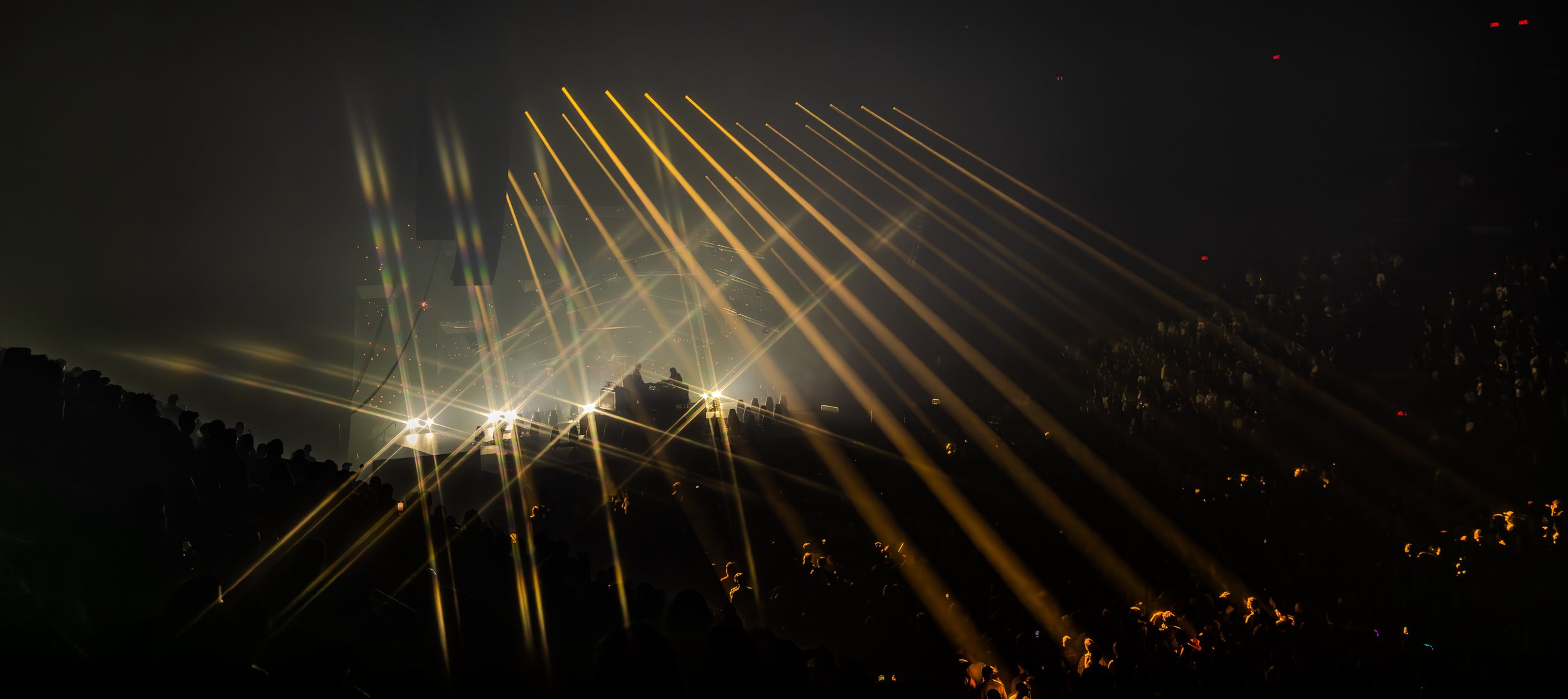 Un concert ou un festival de musique nocturne avec des jeux de lumières jaunes et blanches illuminant la scène et le public silhouetté.